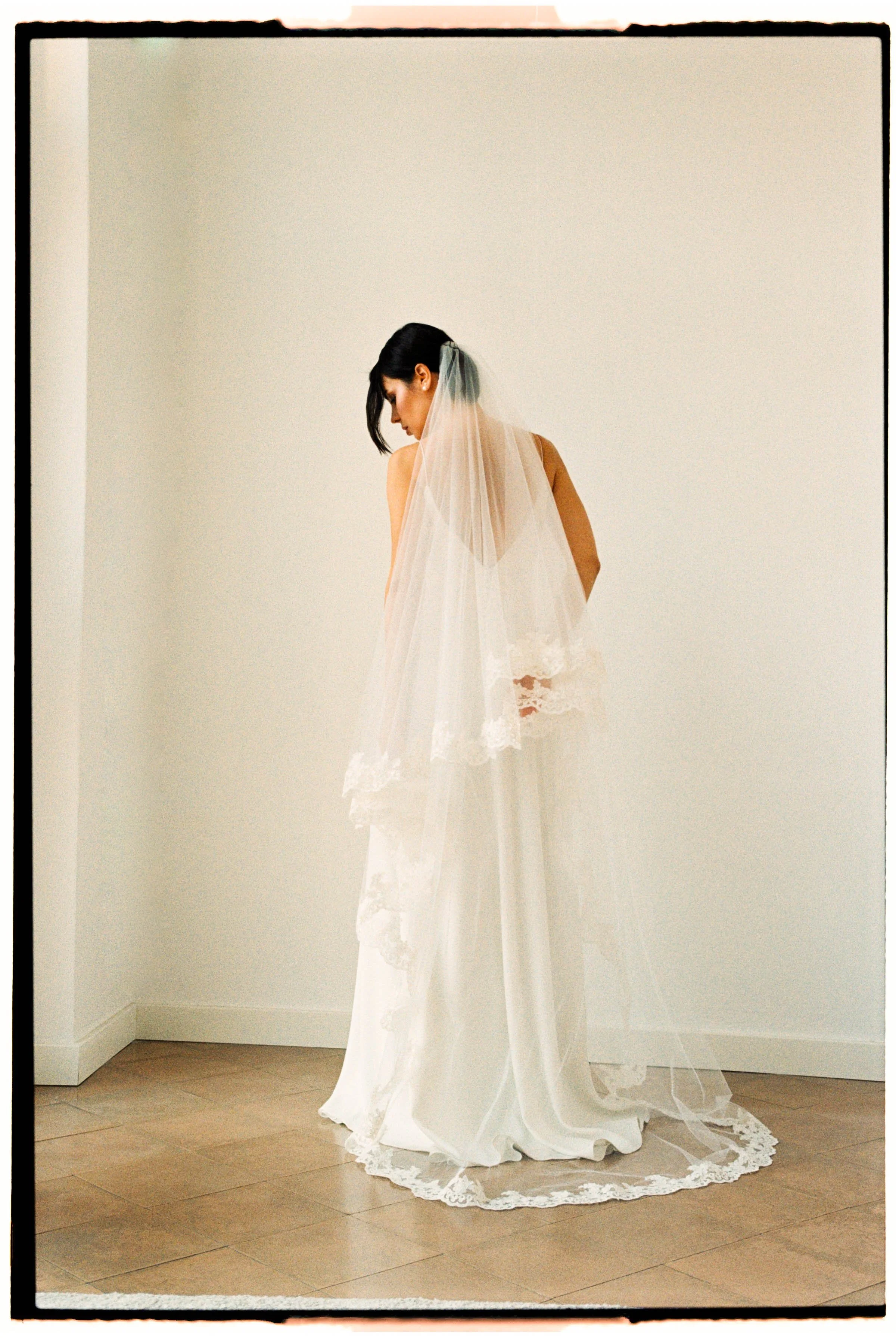 Mujer con vestido de novia, vista de espaldas, con velo largo y encaje, en una habitación con paredes blancas y piso de madera.