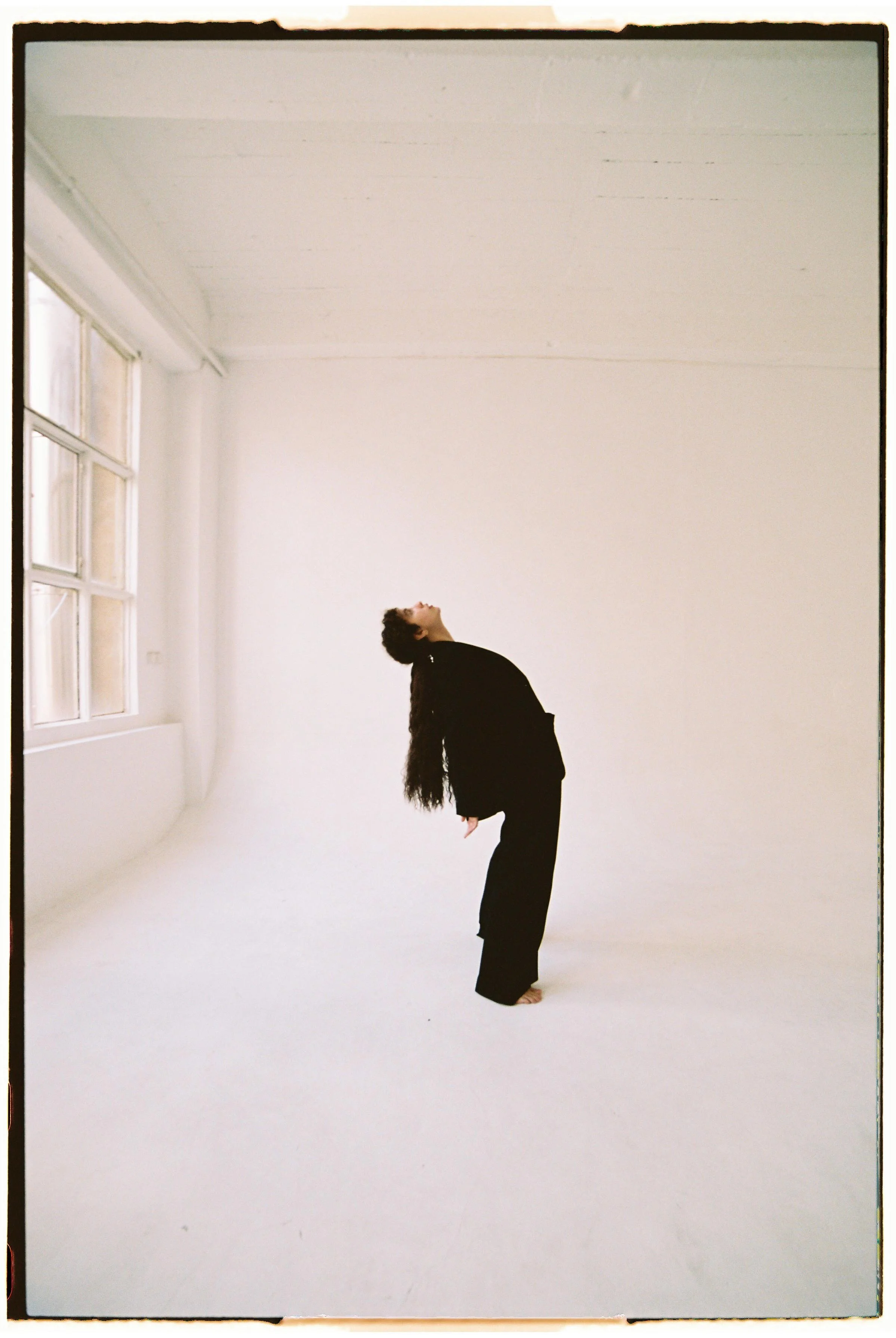 Persona en un espacio blanco y amplio, inclinado hacia atrás, con cabello largo y oscuro, vestida con ropa negra, en una postura de baile o ejercicio.