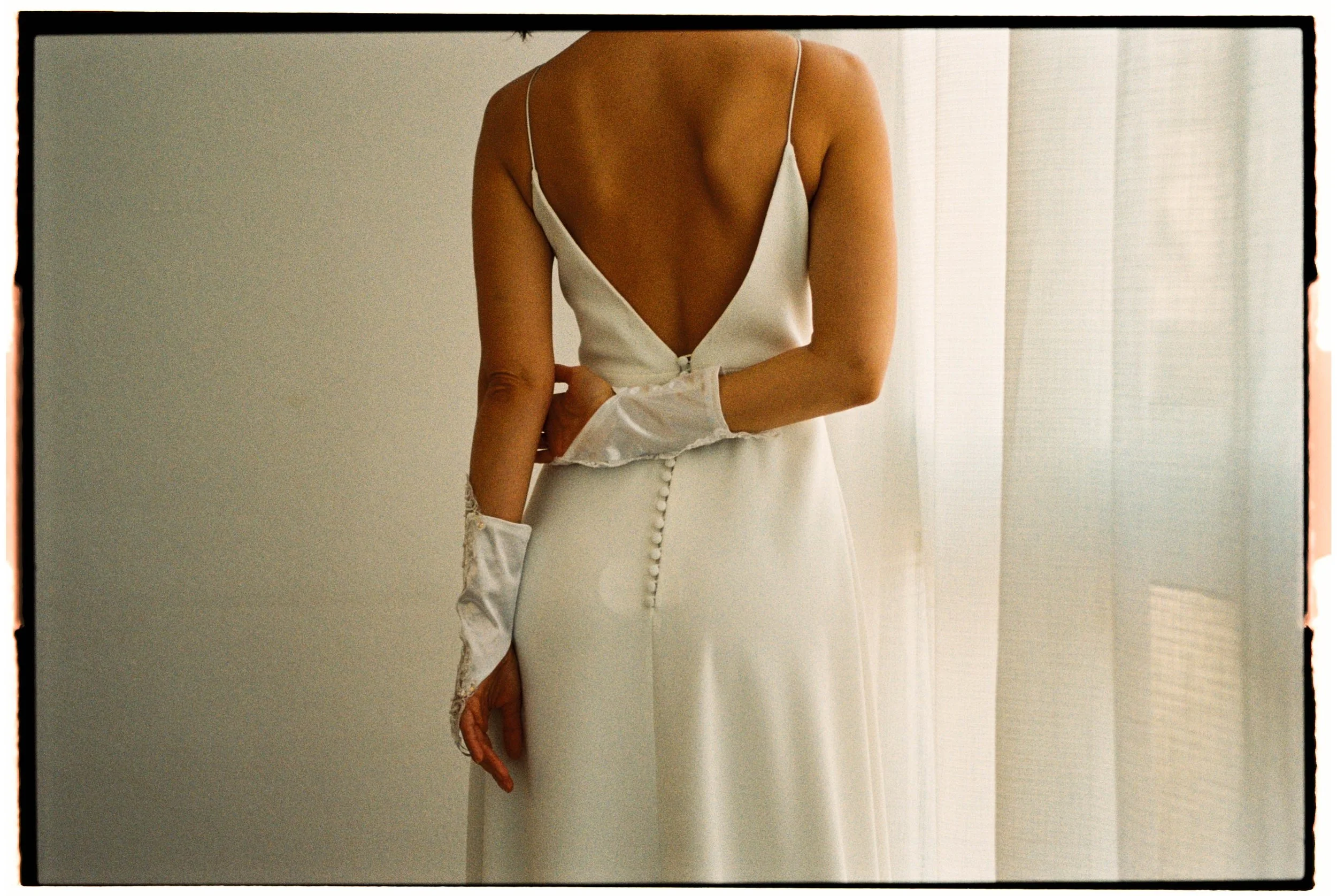 Mujer vista de espaldas con vestido blanco de novia, con escote en V profundo y botones en la parte posterior, sosteniendo un guante largo de encaje.