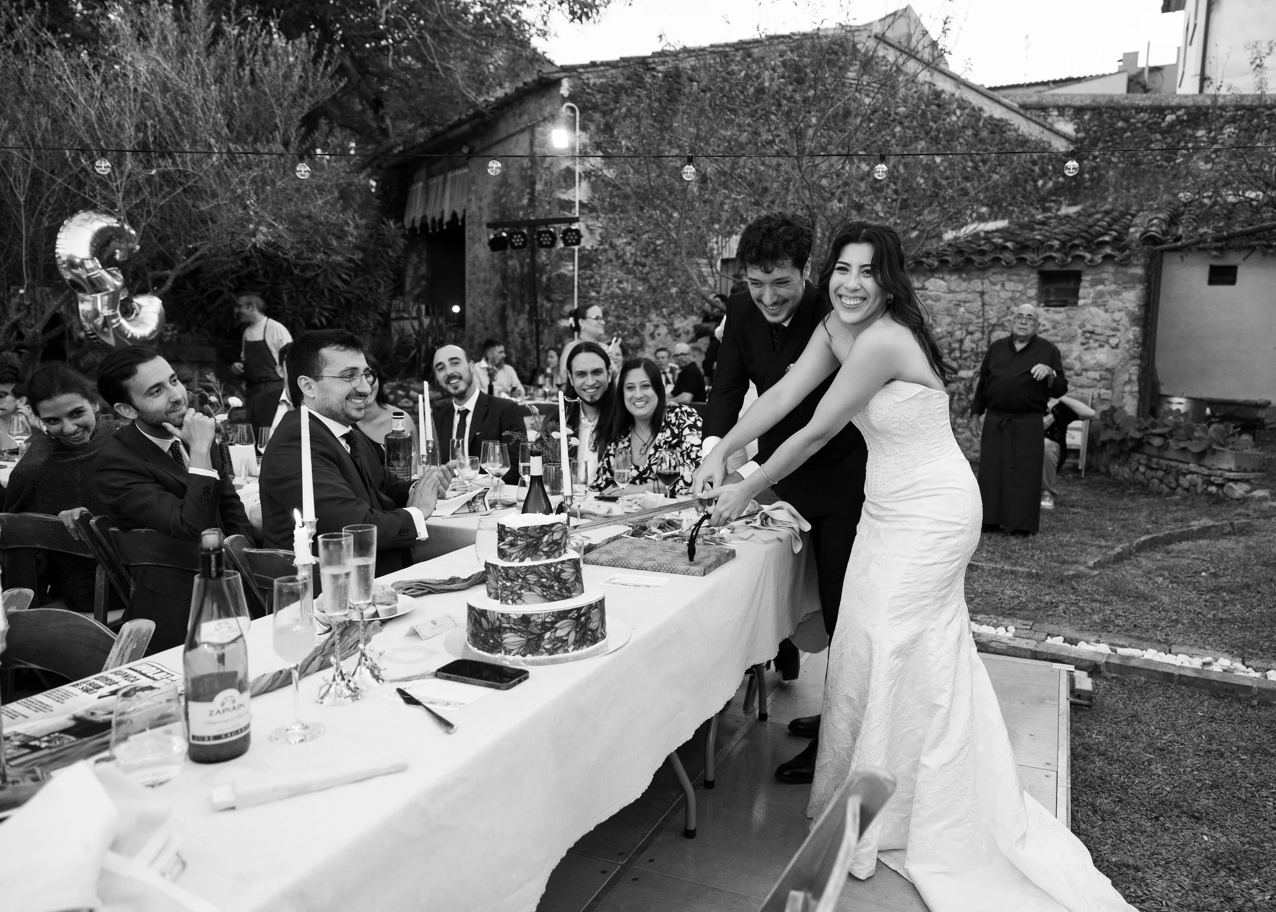 Pareja en vestido de novia y traje en celebración con varios invitados en una mesa larga al aire libre, decorada con velas, en un evento que parece ser una boda, en un espacio exterior con árboles y una casa de fondo.