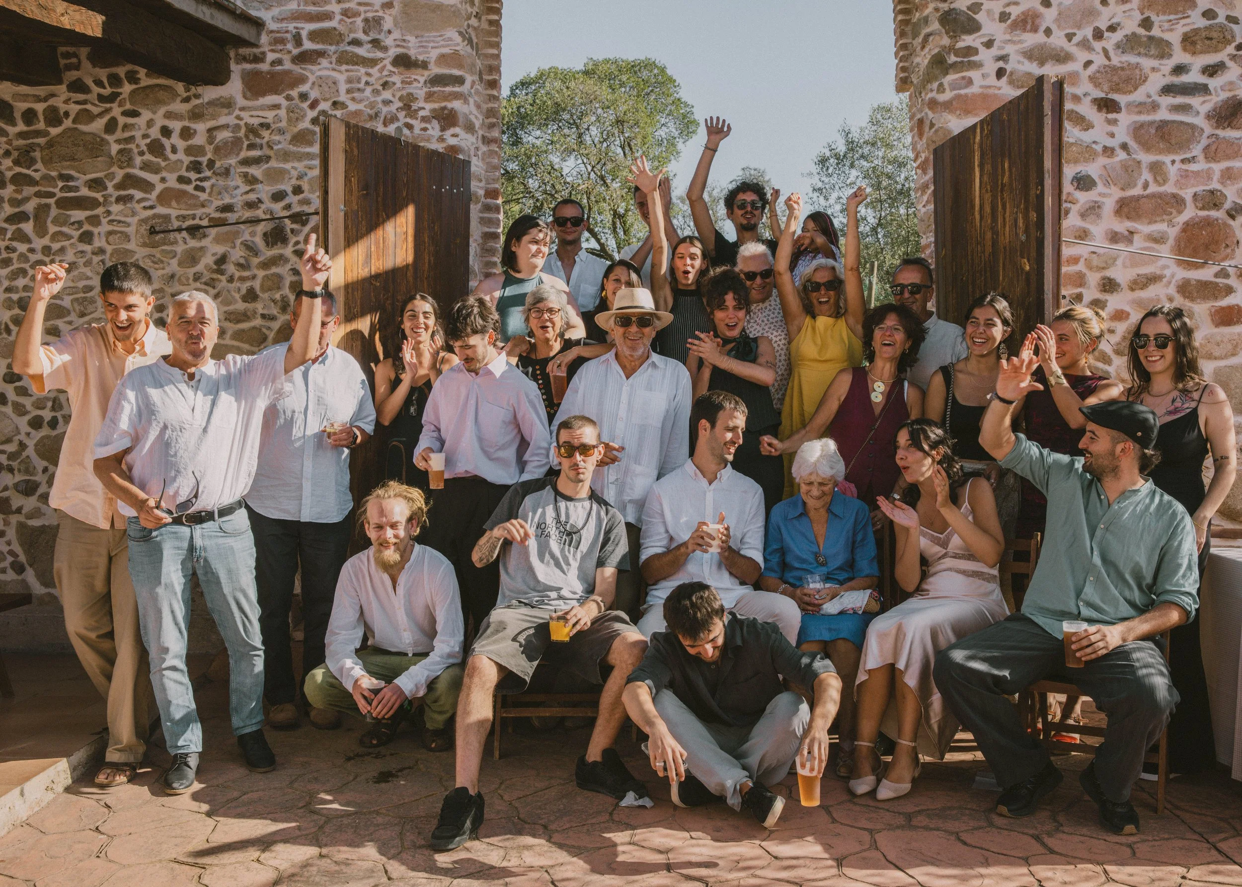 Grupo de personas celebrando, levantando las manos y sonriendo frente a un muro de piedra en un día soleado.