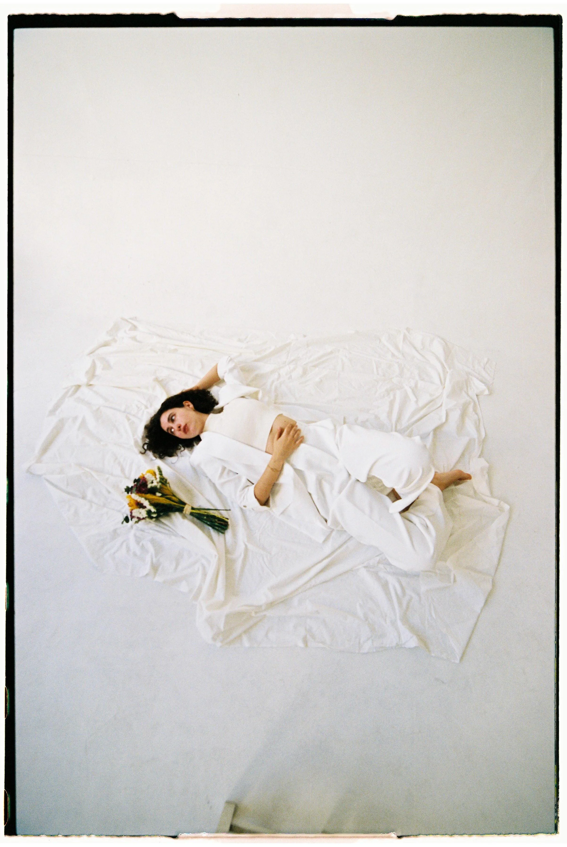 Mujer acostada sobre sábanas blancas con flores en la mano, en una habitación blanca.