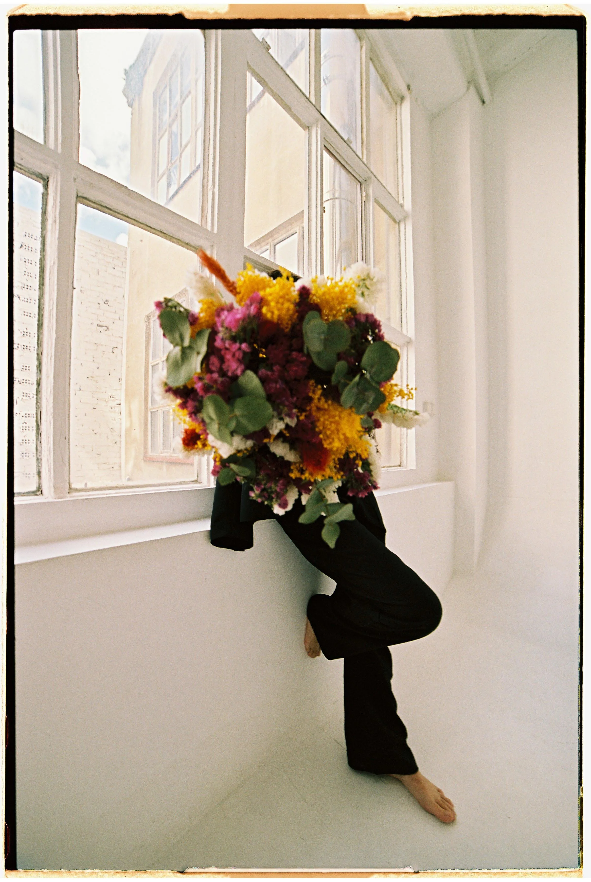 Una persona con pantalones negros, sin zapatos, sostiene un gran ramo de flores frente a una ventana de madera, en un espacio con paredes blancas y luz natural.