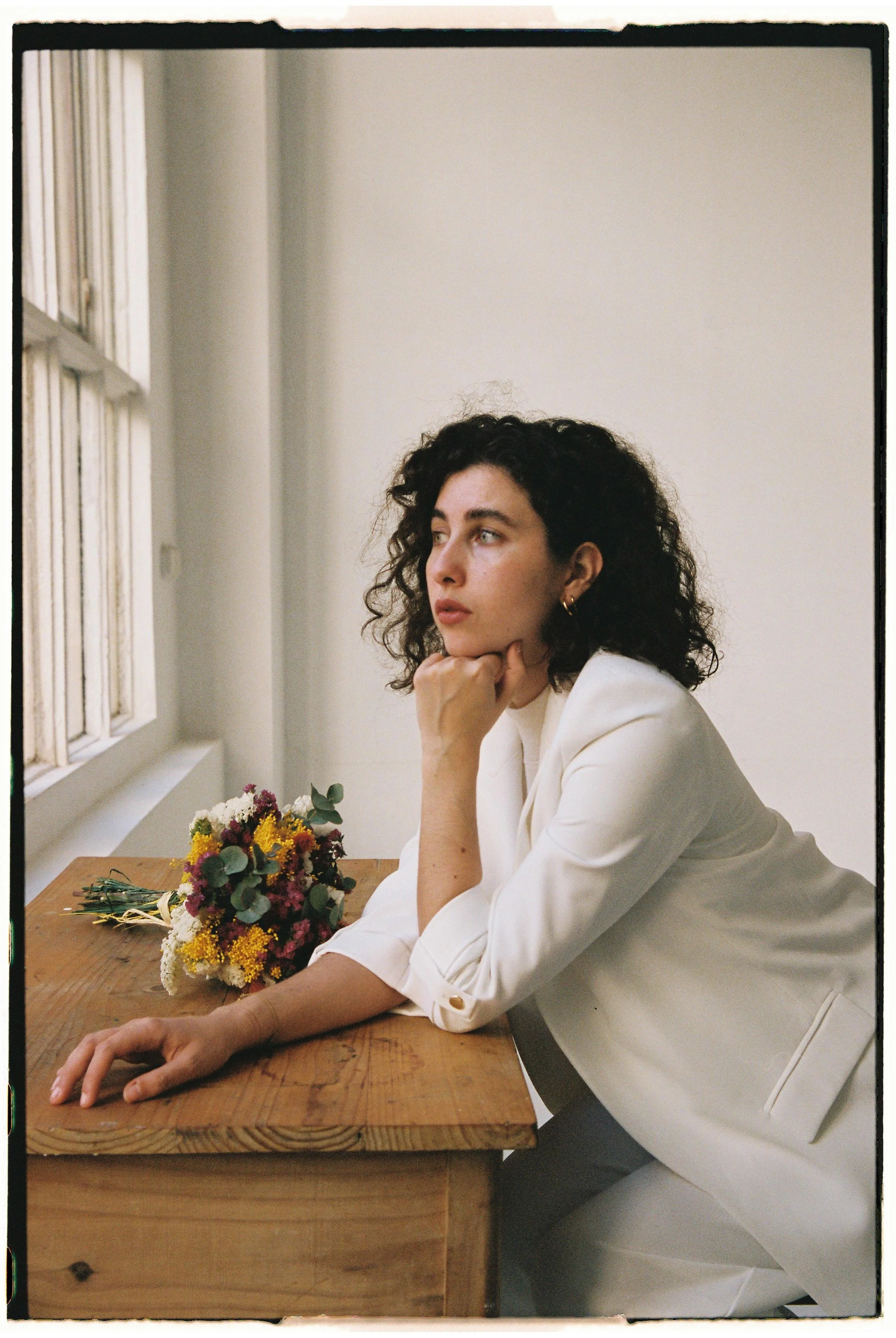 Mujer con cabello rizado oscuro, vestida con saco blanco, sentada en una mesa de madera con un ramo de flores sobre ella, mirando hacia la izquierda, en un ambiente interior con ventanas y paredes blancas.
