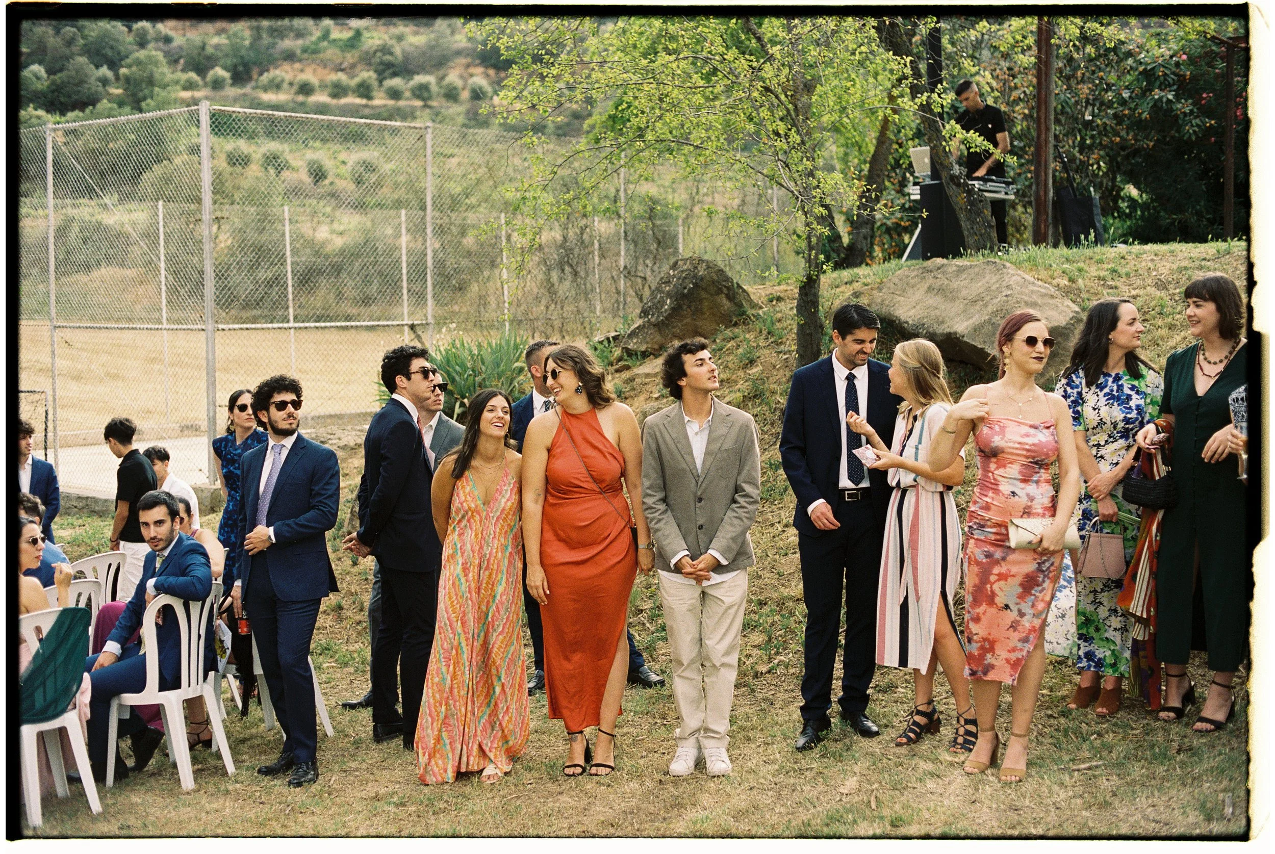Grupo de personas en un evento al aire libre, algunas sentadas y otras de pie, con vestimenta formal y elegante, en un entorno natural con árboles, montañas y una cancha de tenis en el fondo. Hay un DJ en la esquina del fondo tocando música.