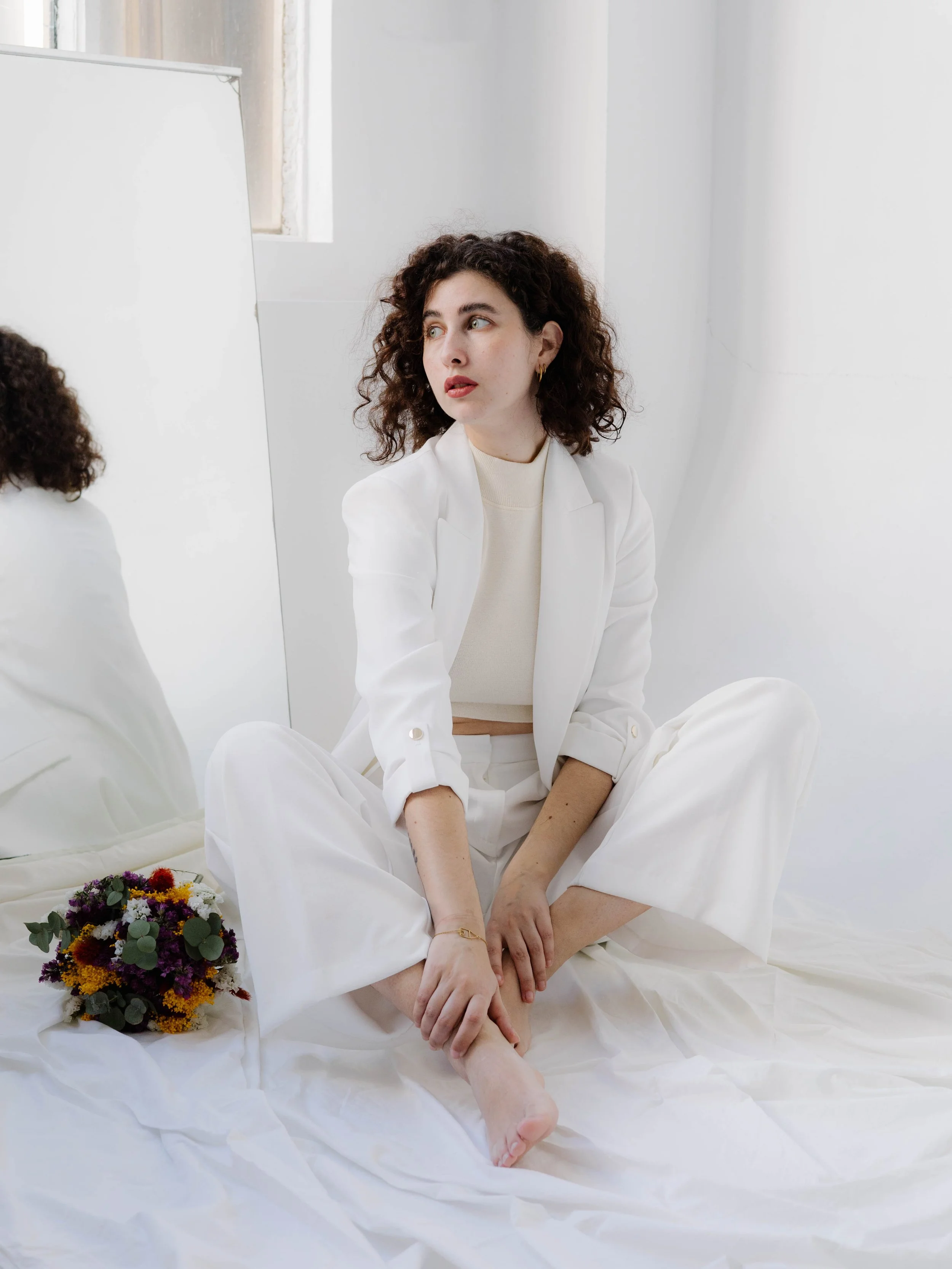 Mujer de cabello rizado sentado en un suelo cubierto con sábanas blancas, con un ramo de flores junto a ella y un espejo en la pared. Ella viste un conjunto blanco y mira hacia la izquierda.