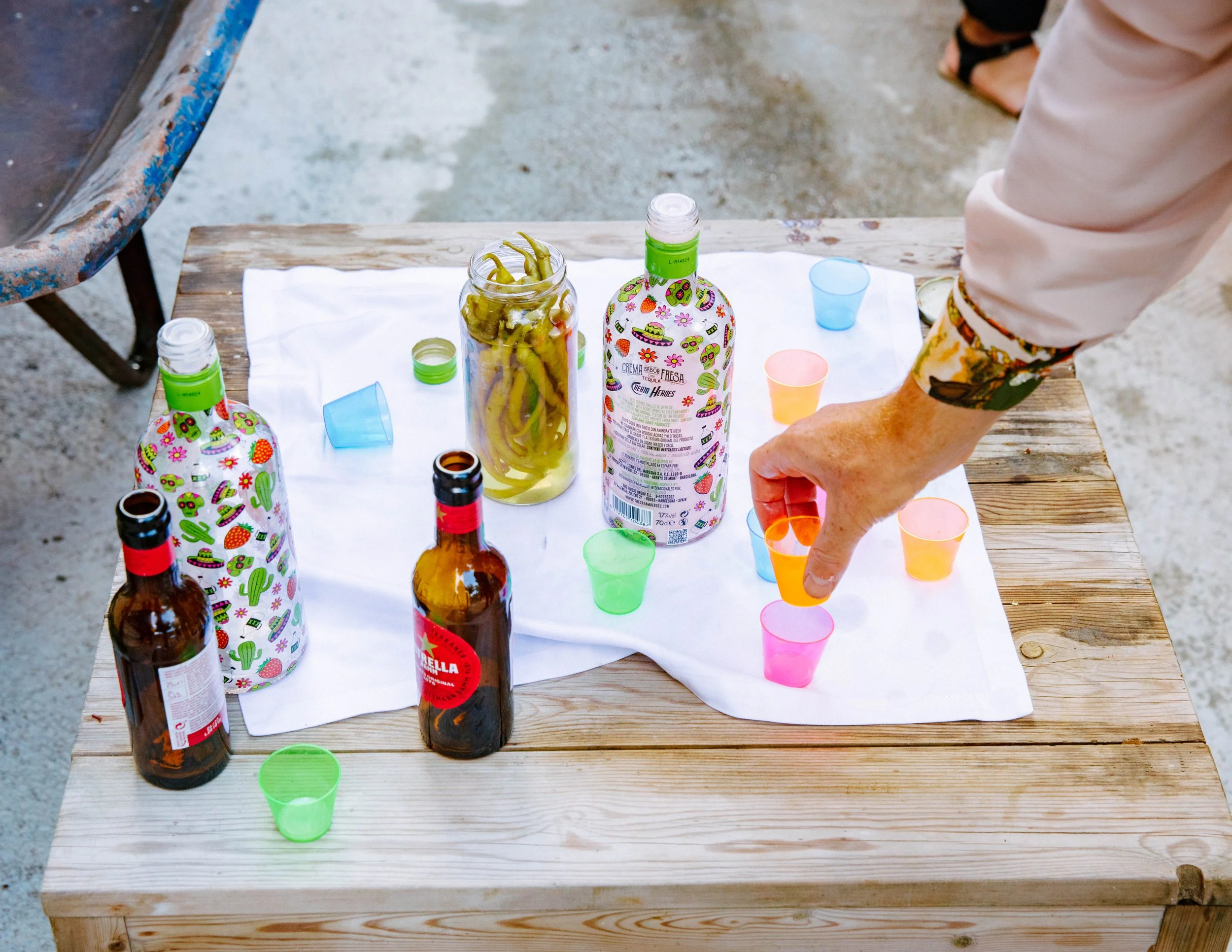 Diversas botellas de cerveza y vasos de colores en una mesa de madera, con una botella decorada con imágenes de cactus, flores y calaveras.