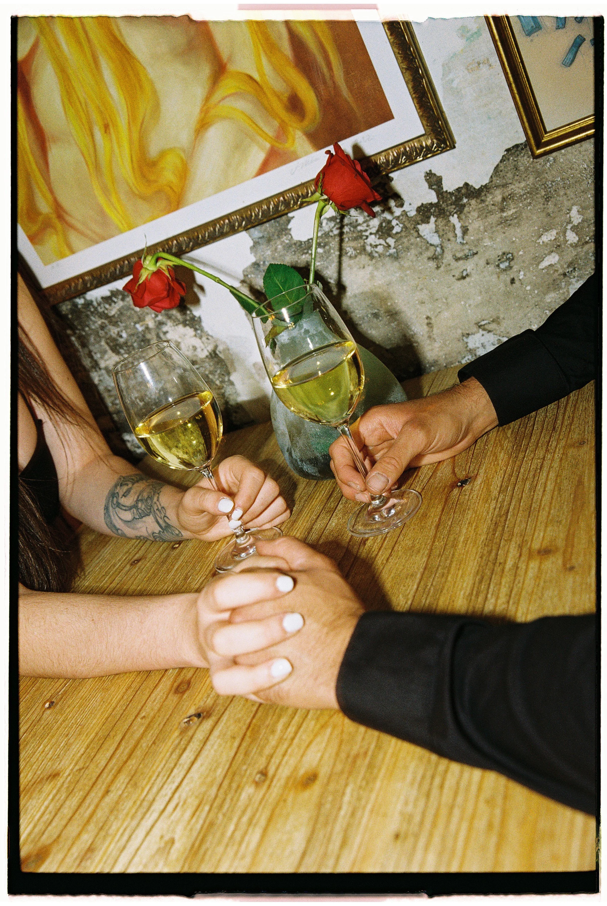 Dos personas con uñas pintadas de blanco brindando con copas de vino en una mesa de madera, con un florero con dos rosas rojas y pinturas en el fondo.