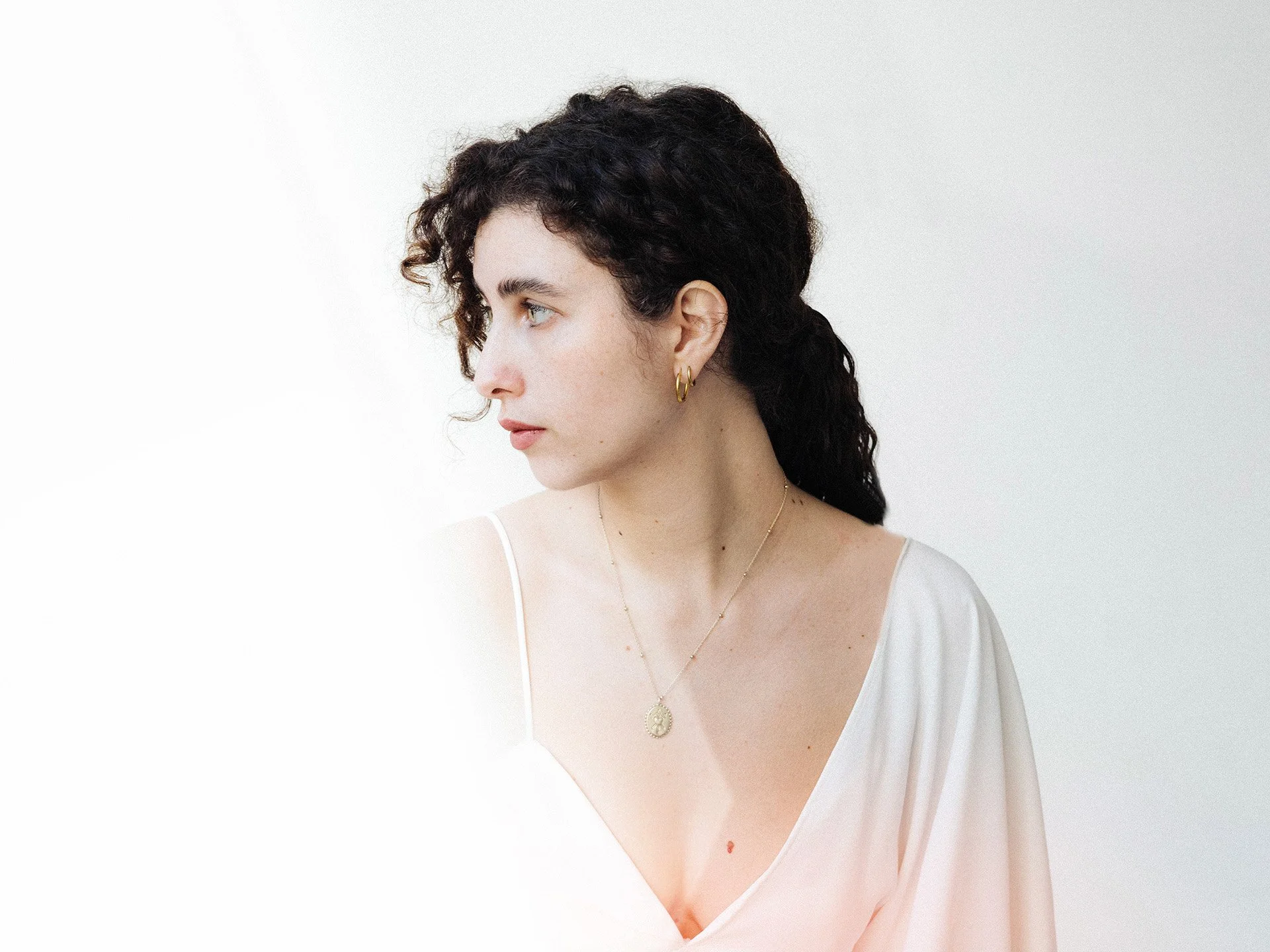 Retrato de mujer con cabello rizado oscuro mirando hacia la izquierda, usando pendientes de aro y collar de oro, con blusa blanca, en fondo blanco.