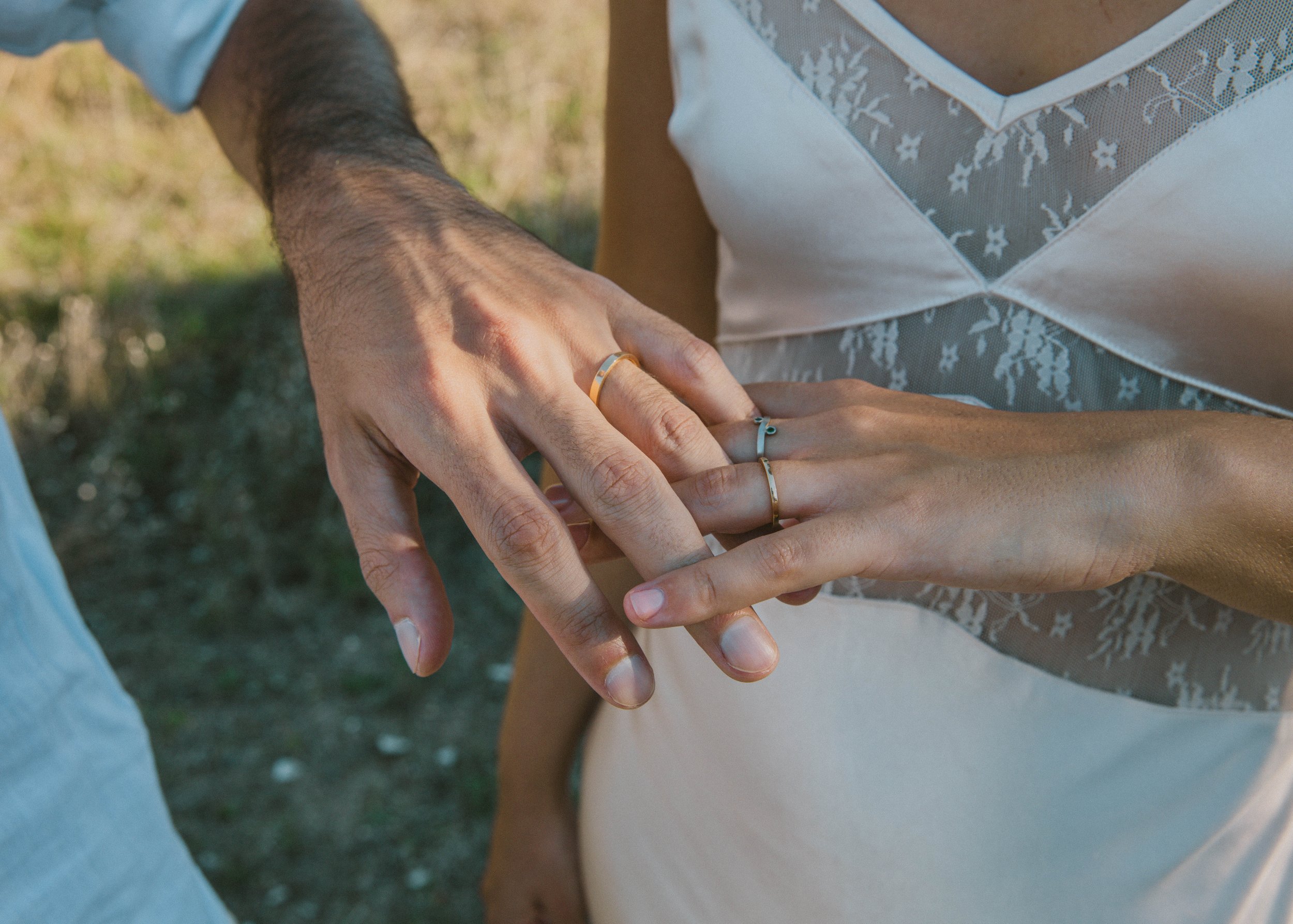 Primer plano de las manos de una pareja con anillos, tocándose los dedos. La mujer lleva un vestido blanco con detalles de encaje y la mano del hombre tiene un reloj. Fondo natural al aire libre.