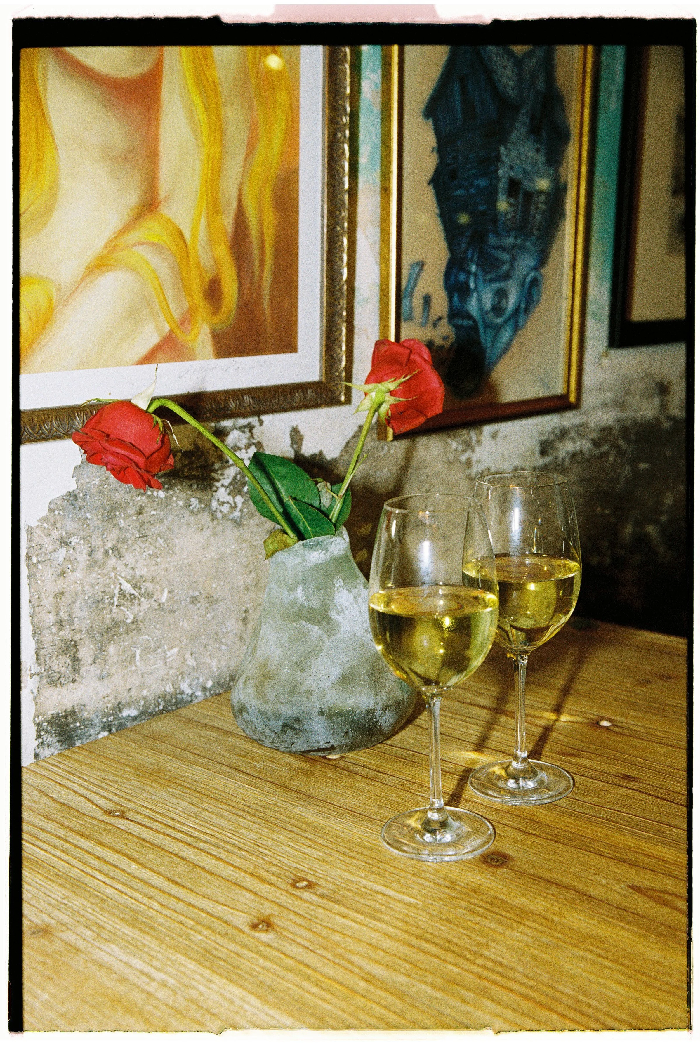 Dos copas de vino blanco sobre una mesa de madera, junto a un florero con dos rosas rojas, en un fondo con cuadros y pared de cemento.