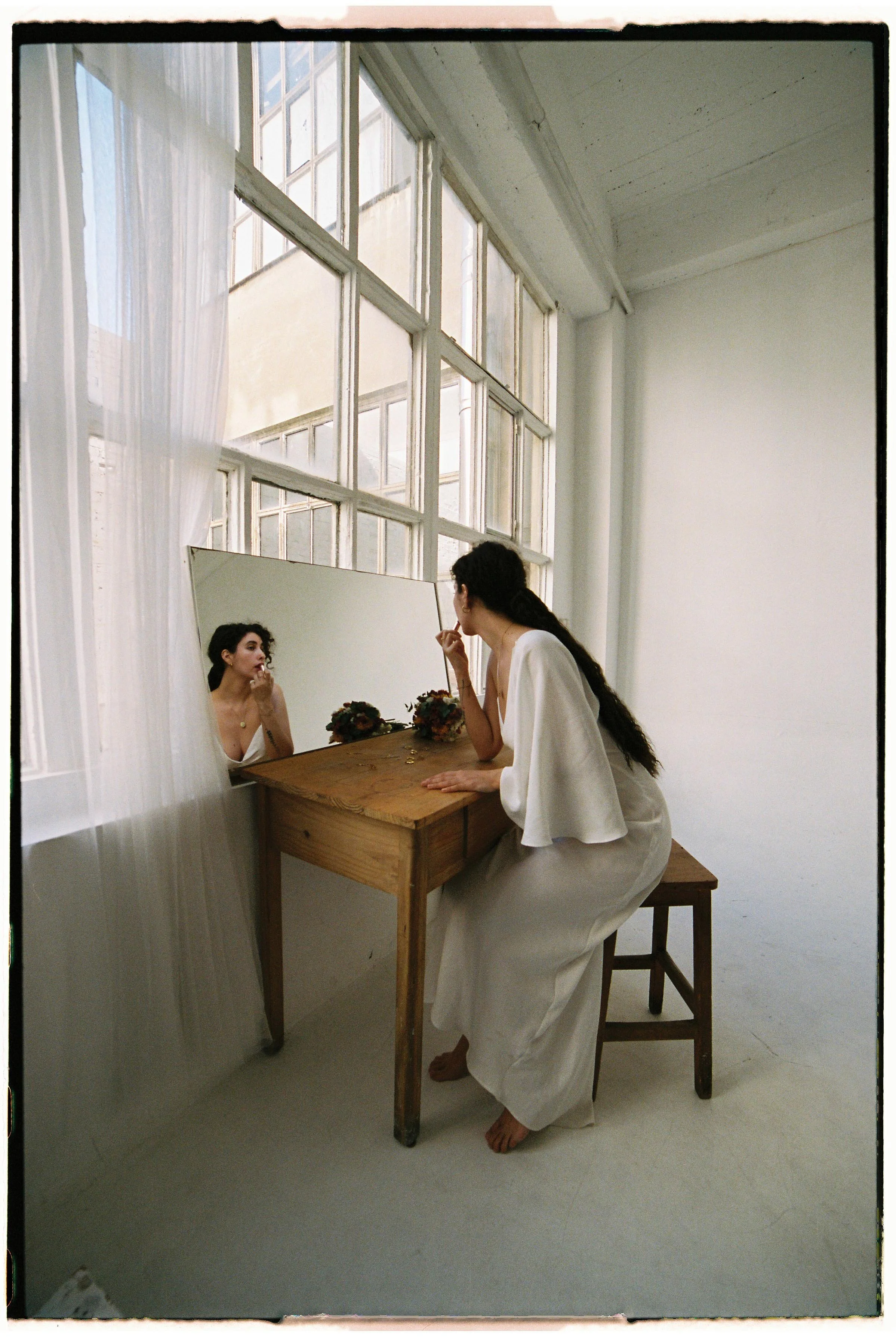 Mujer con vestido blanco sentado en una silla frente a un espejo, arreglándose el cabello en una habitación luminosa con ventanas grandes y cortinas blancas.