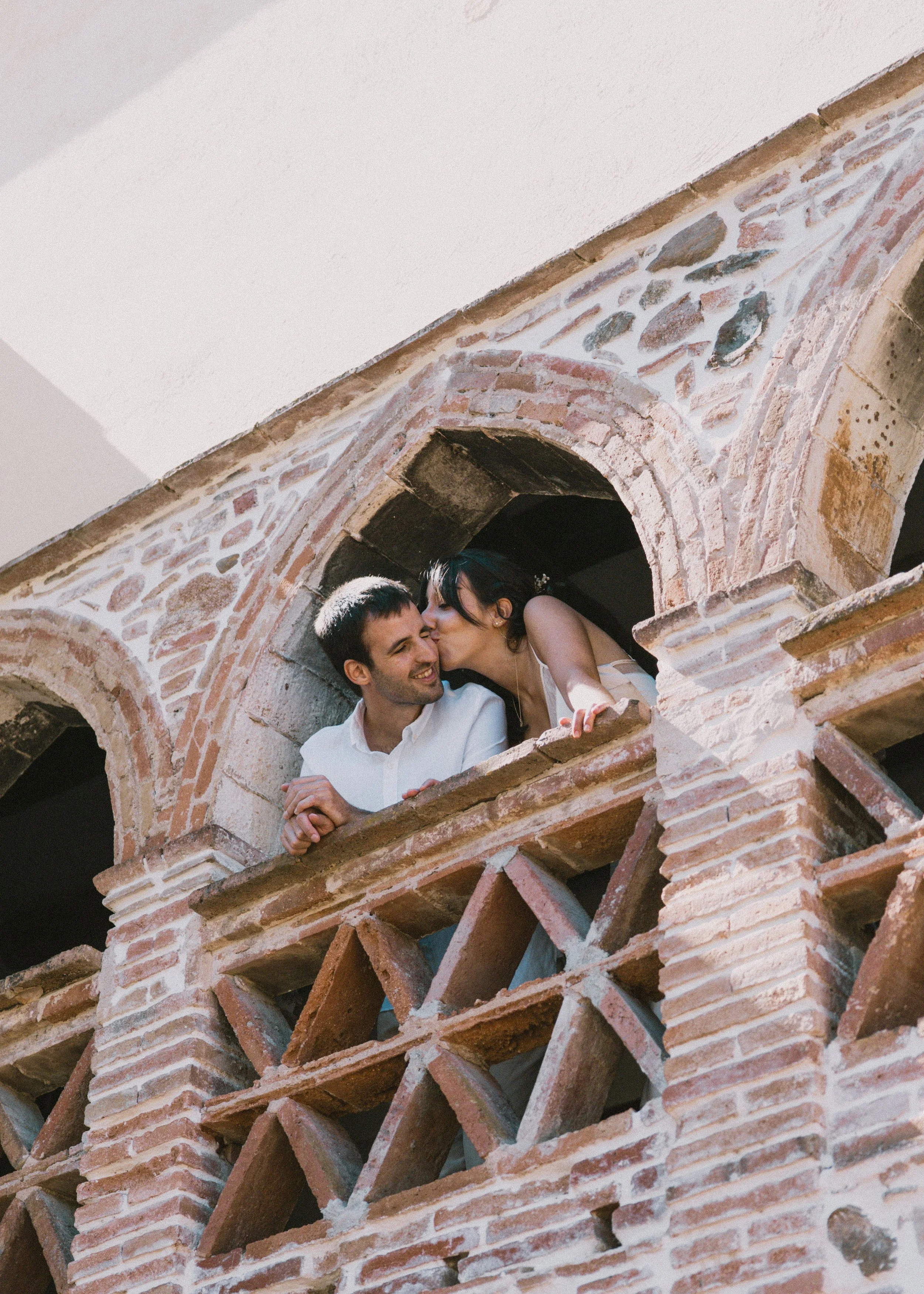 Una pareja en un balcón de ladrillos, ella le da un beso en la mejilla a él. Ambos sonríen, en un ambiente de arquitectura antigua con arcos y ventanas de piedra.