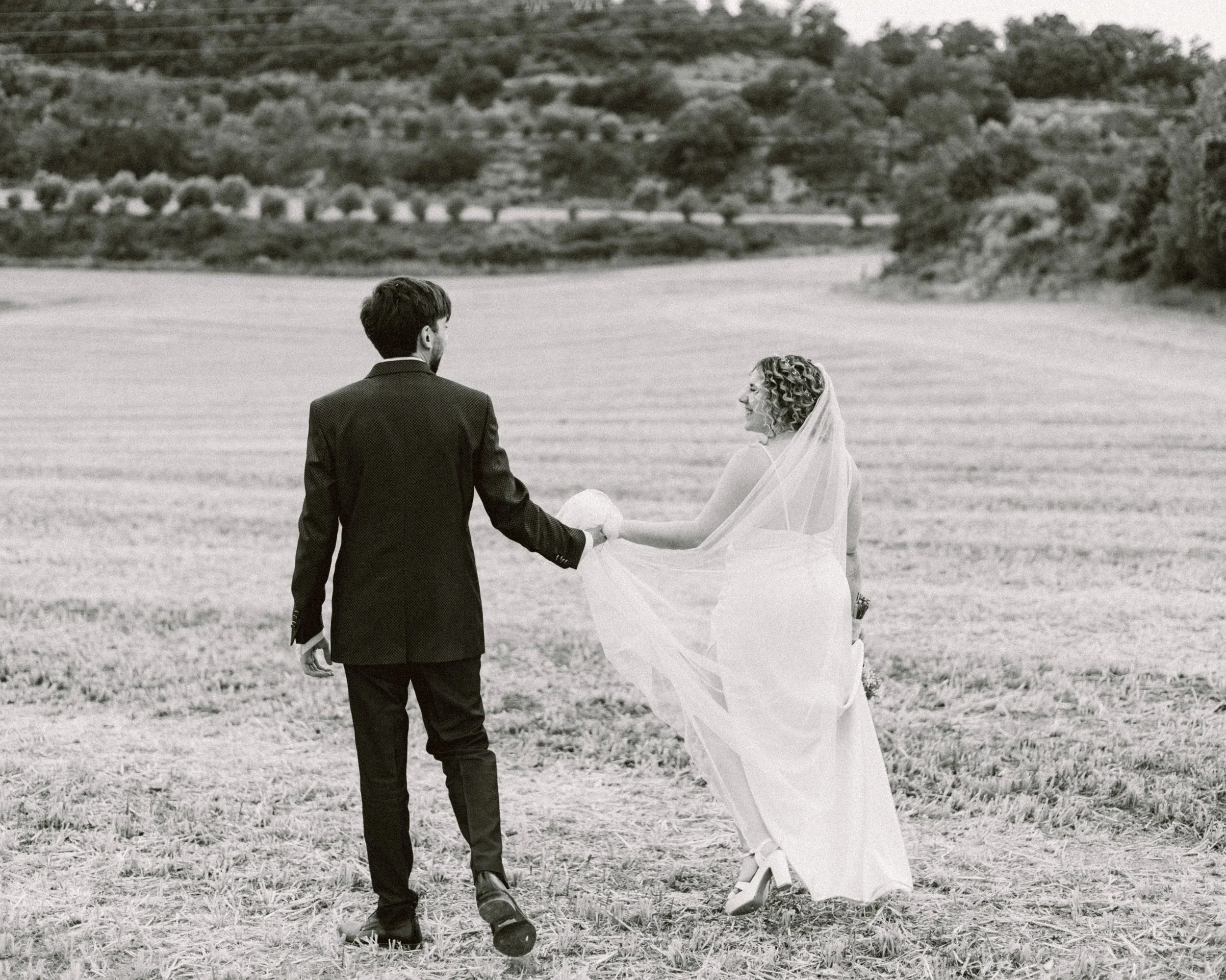 Pareja de novios en un campo abierto, caminando juntos, la mujer con vestido de novia y velo, el hombre con traje oscuro, en un entorno natural.
