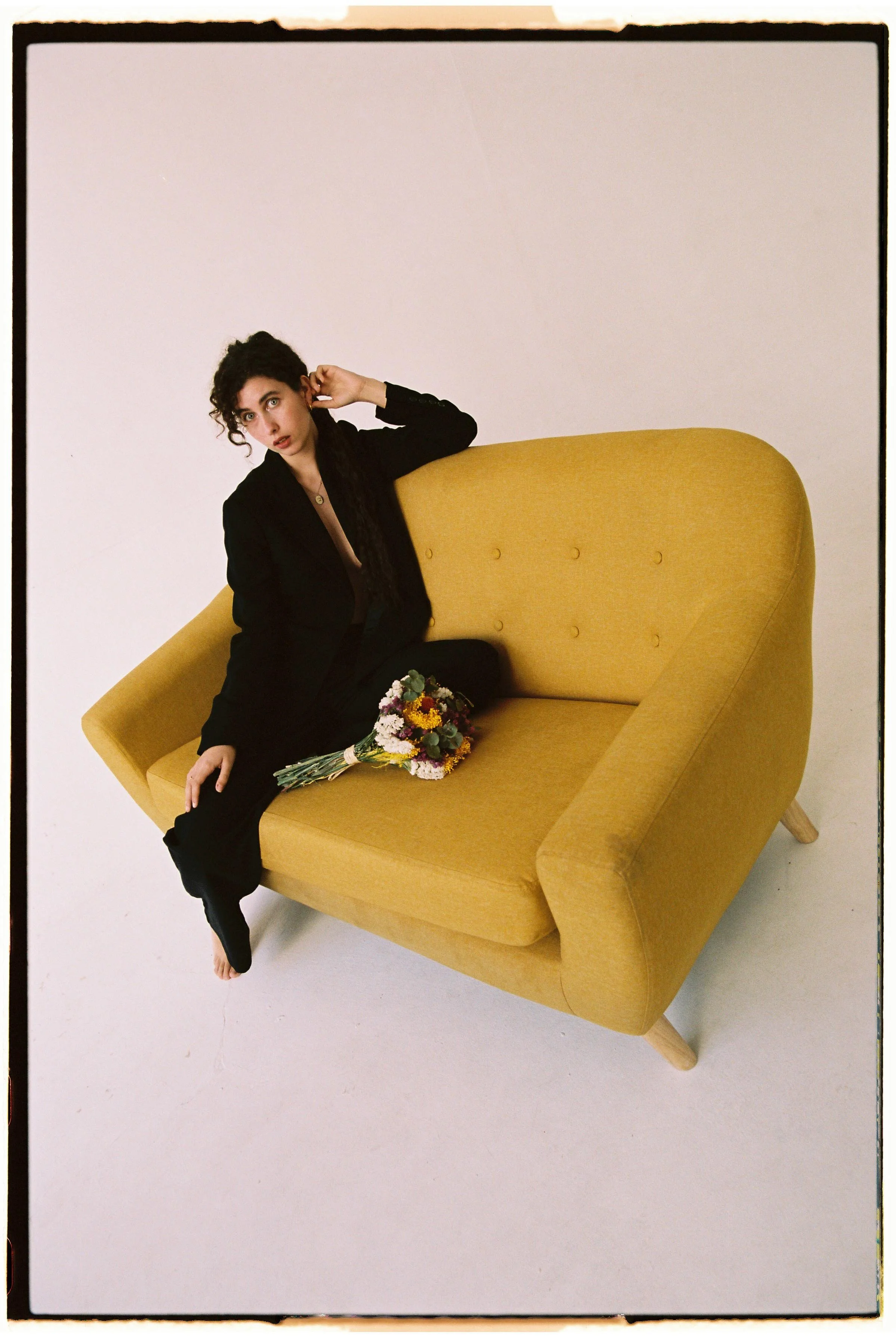 Una mujer sentada en un sillón amarillo, sosteniendo un ramo de flores, vestida con ropa negra, en un fondo blanco.