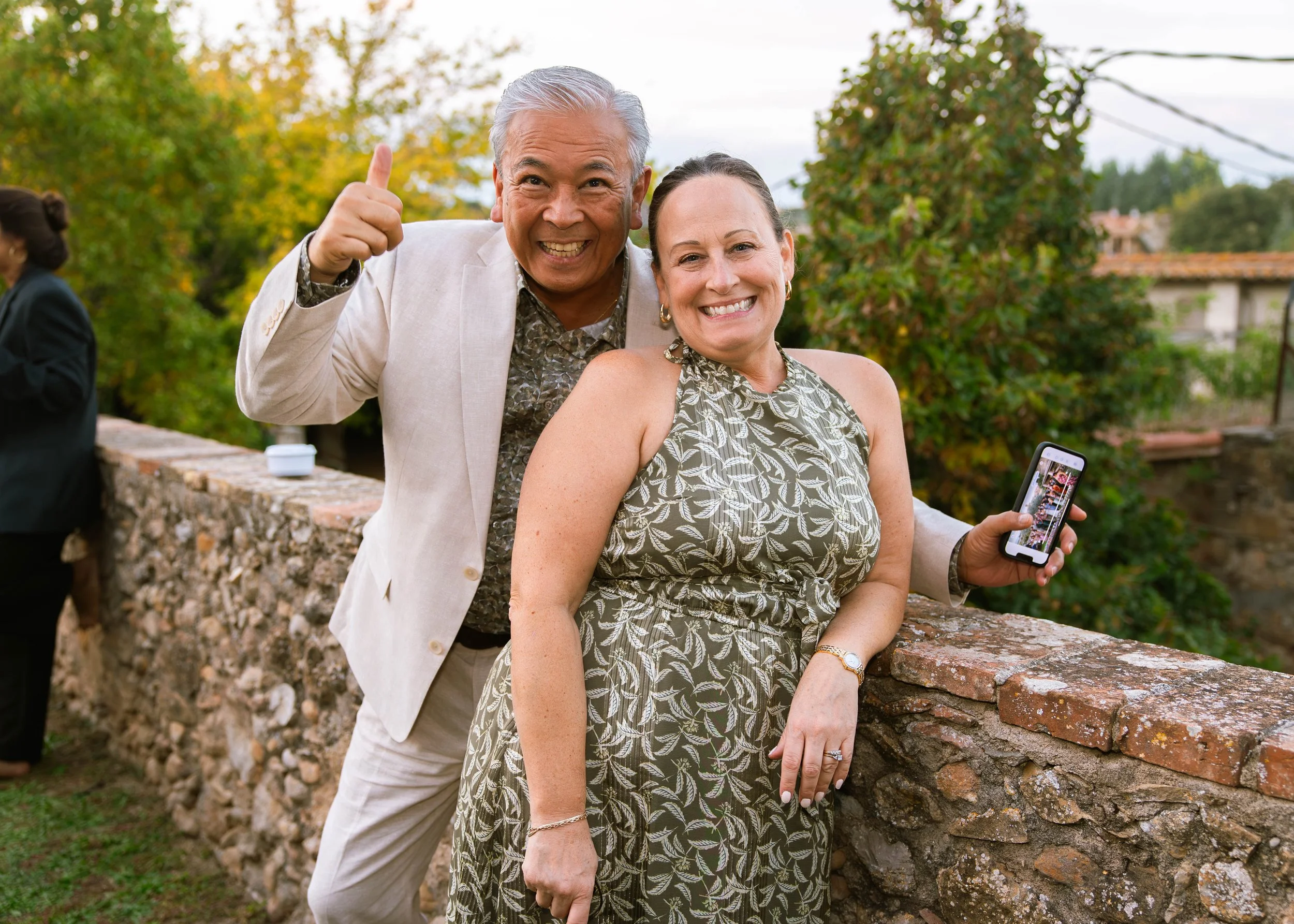 Hombre mayor con piel oscura y cabello gris, vestido con traje claro, sonriente y saludando, junto a mujer con piel clara y cabello oscuro, vistiendo vestido con estampado de hojas, sonriendo y sosteniendo un teléfono móvil, en un entorno exterior co