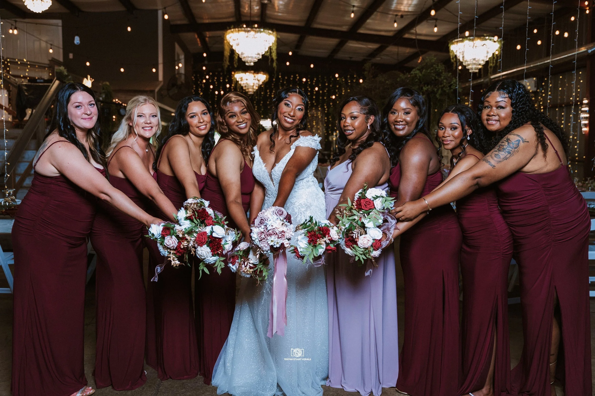 A bride in a white wedding dress standing among eight bridesmaids in wine-colored dresses, all holding bouquets and posing together at a wedding reception with dim lighting and hanging chandeliers.