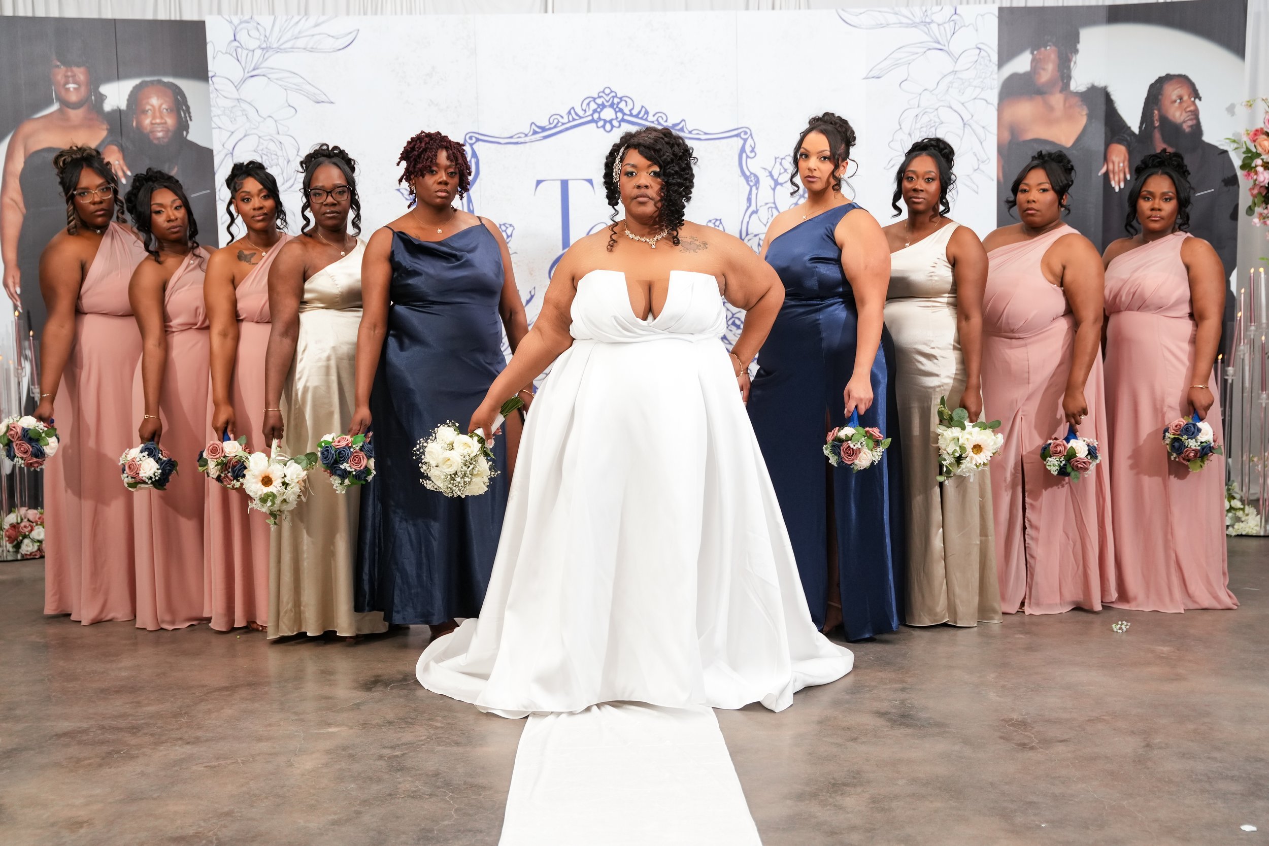 A group of women in formal dresses standing together at a wedding or special event, with a central woman in a white gown, holding bouquets of flowers, posing for a group photo indoors.