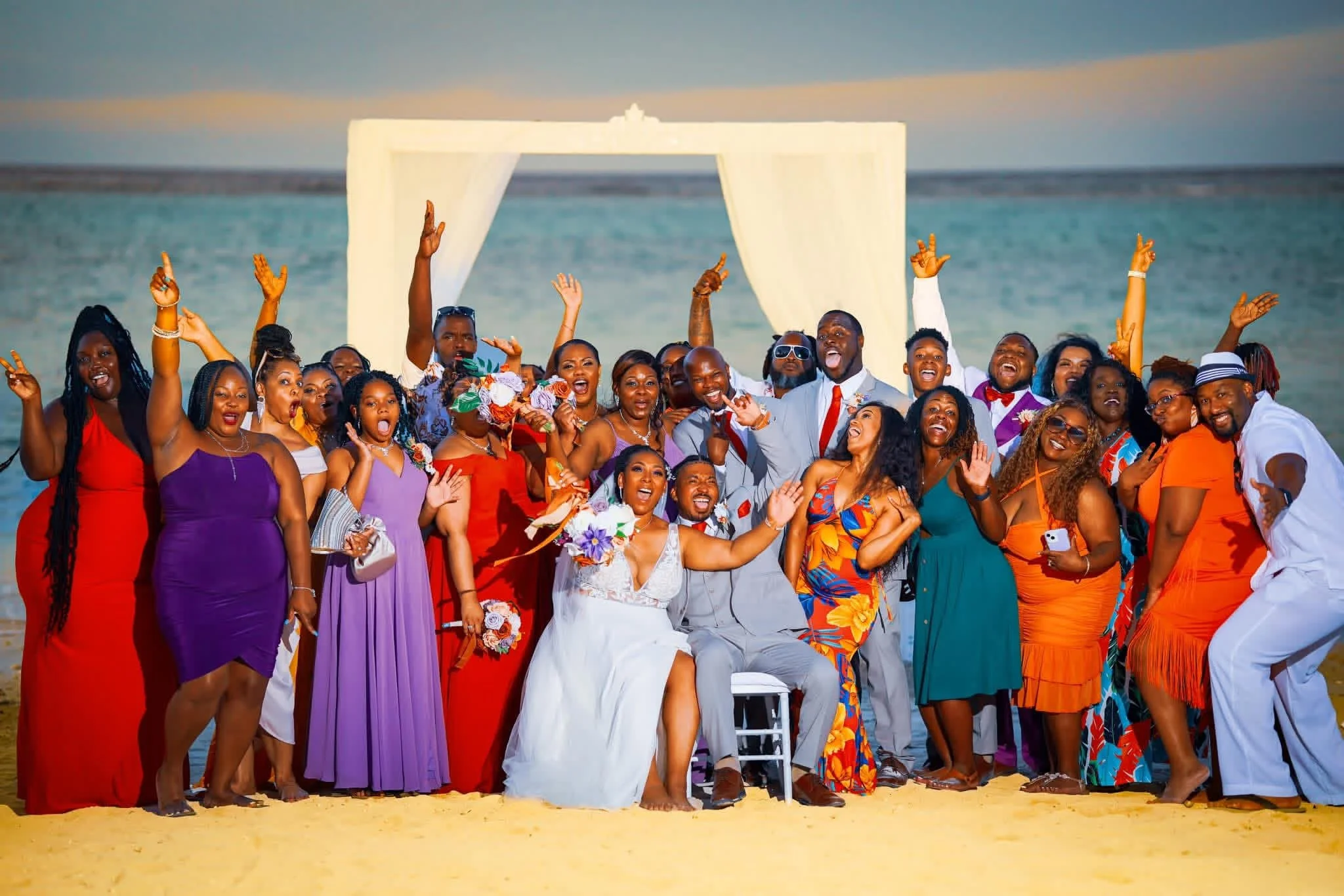 A large group of people dressed in colorful beachwear celebrating on a sandy beach with an ocean backdrop and a white canopy structure.