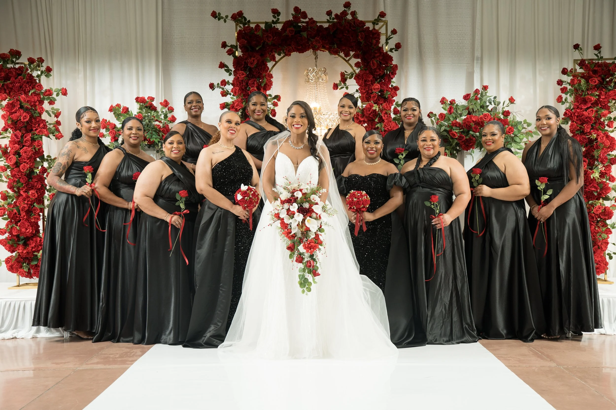 A bride in a white wedding dress holding a bouquet standing in front of a group of women in black dresses at a wedding celebration with red roses and floral decorations