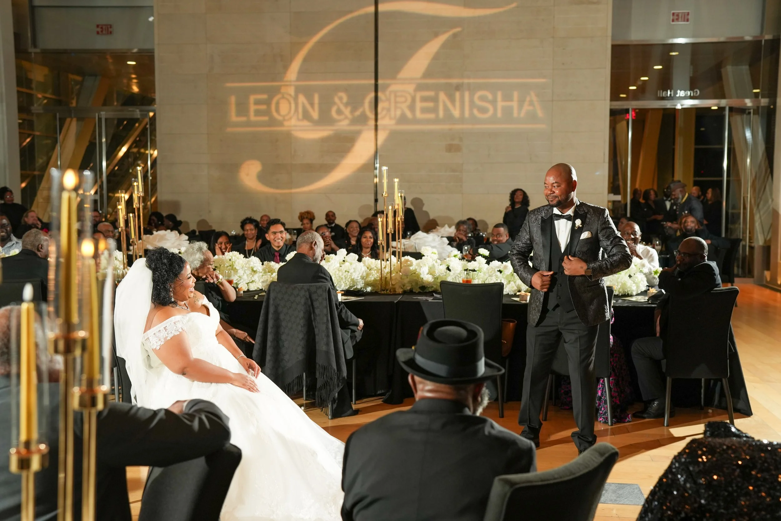 A bride in a white wedding dress sitting at a banquet table, smiling and looking at a man in a tuxedo standing nearby. Guests are seated around the table, and the setting appears to be a wedding reception in a large, modern hall with a projection on the wall that reads 'LEON & GRENISHA'.