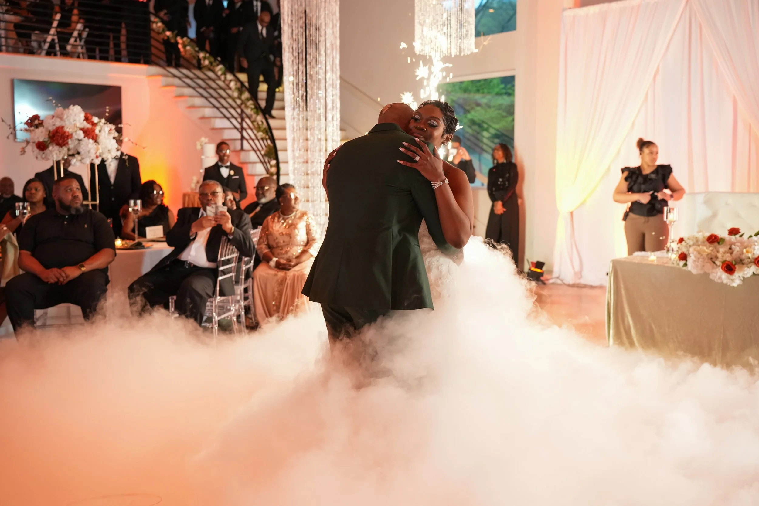 A bride and groom dancing closely at their wedding reception, surrounded by seated guests, floral decorations, and elegant decor with draped curtains and chandeliers.