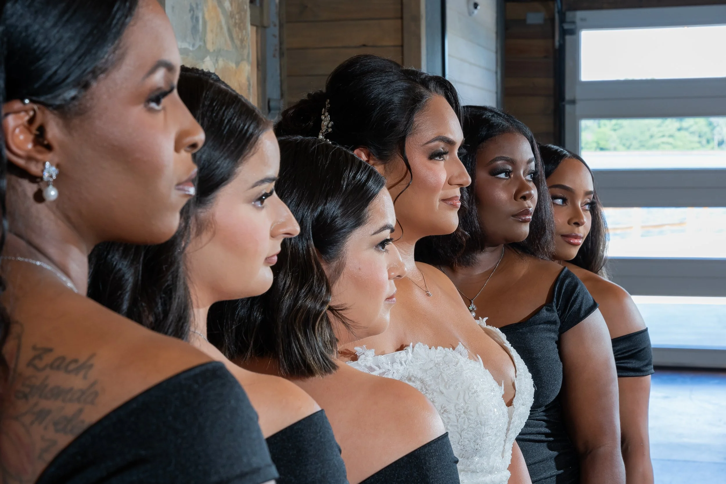 Side profile of seven women dressed in formal attire, lined up and facing to the right, with a background of wooden walls and a large window showing a clear outdoor view.