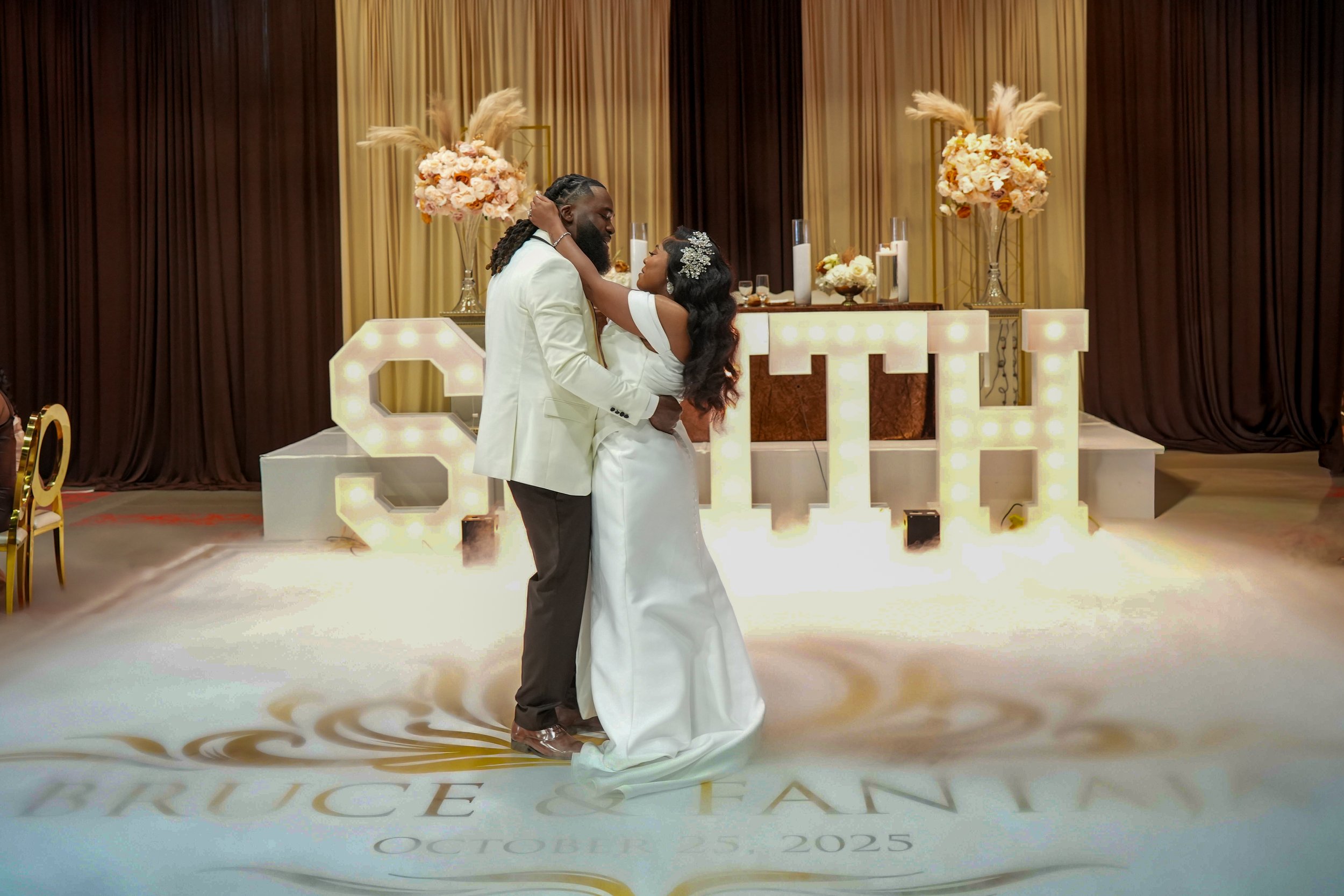A newlywed couple sharing a dance at their wedding reception, with a decorated backdrop and large illuminated 'LOVE' sign.