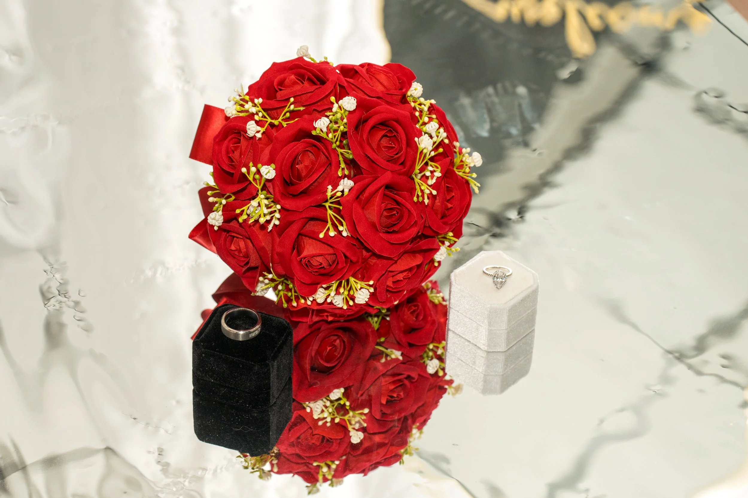 A bouquet of red roses with small white flowers and green foliage, placed on a reflective surface. Nearby are two rings, one silver and one black, and a small white jewelry box. The background has a smooth silver surface.