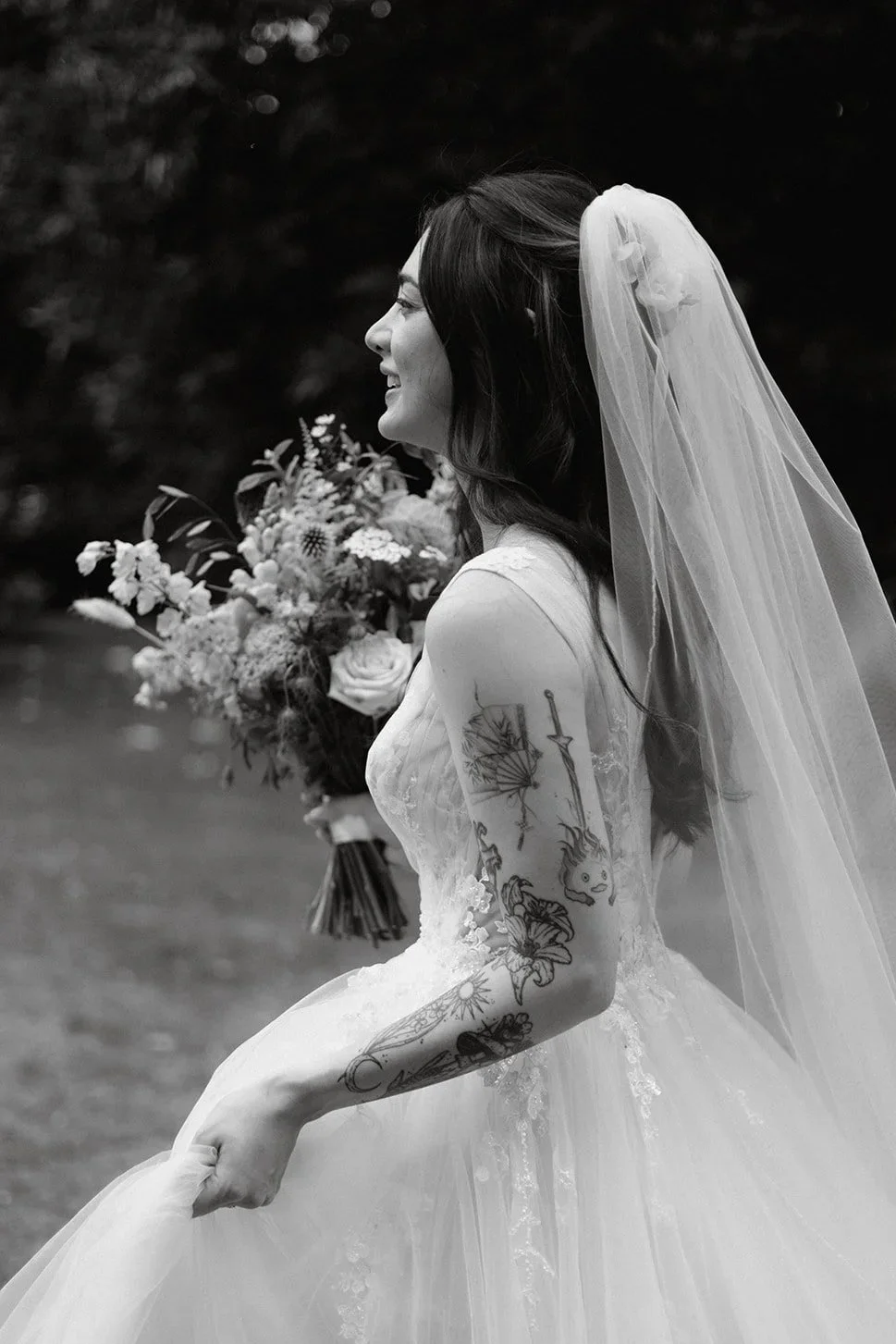 A bride in a wedding dress with a veil, holding a bouquet, standing outdoors with a background of trees, smiling in profile.