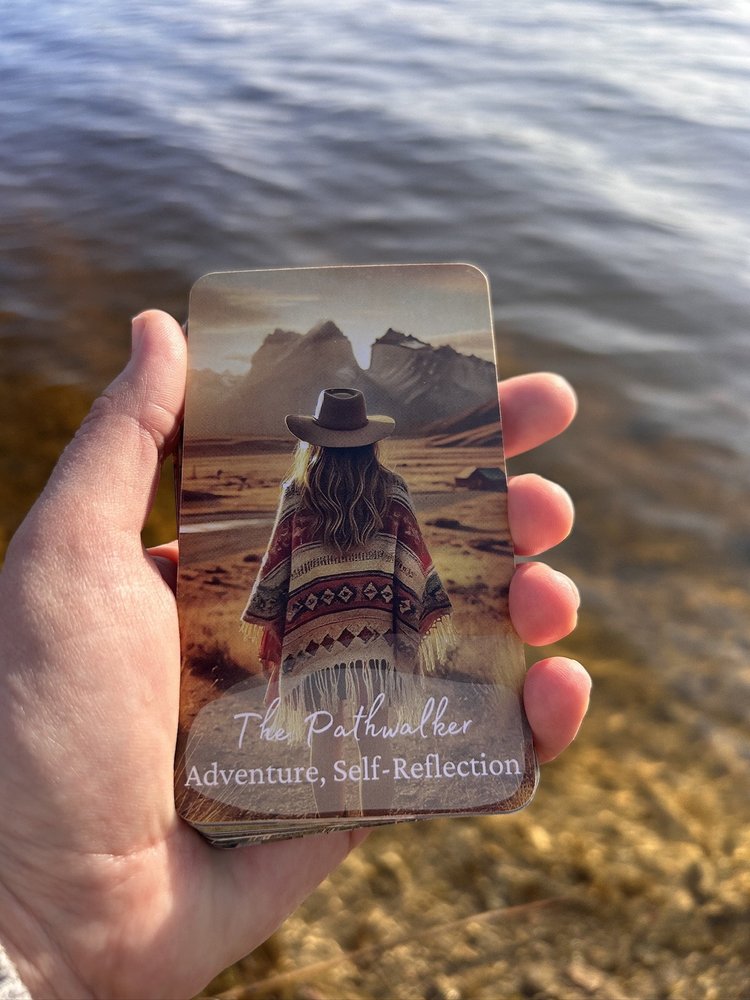 A hand holding The Pathwalker card from Boho Wisdom Oracle before the water of a quiet lake.