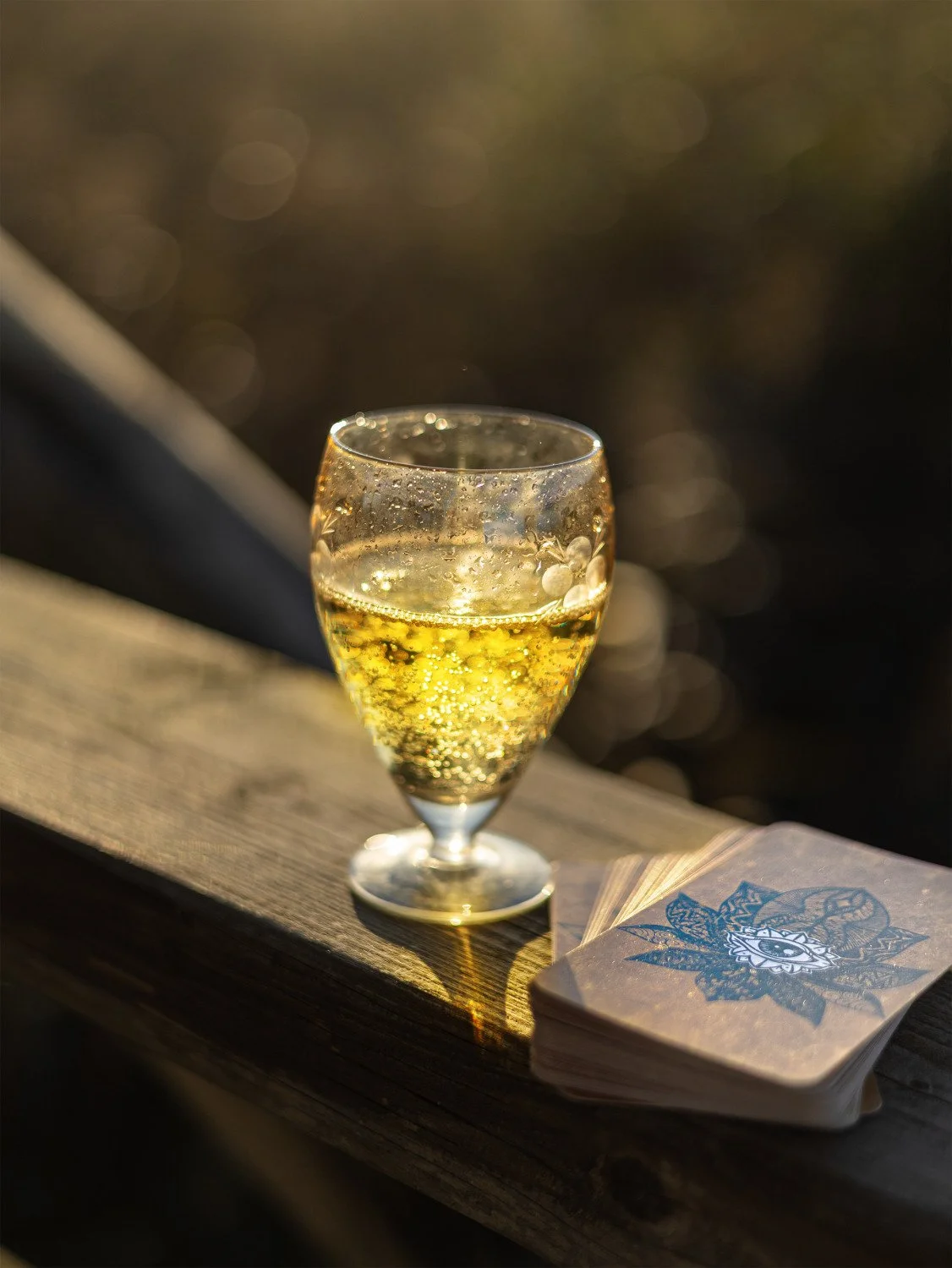 Glass of sparkling drink beside a brown oracle card deck on a sunlit wooden railing.
