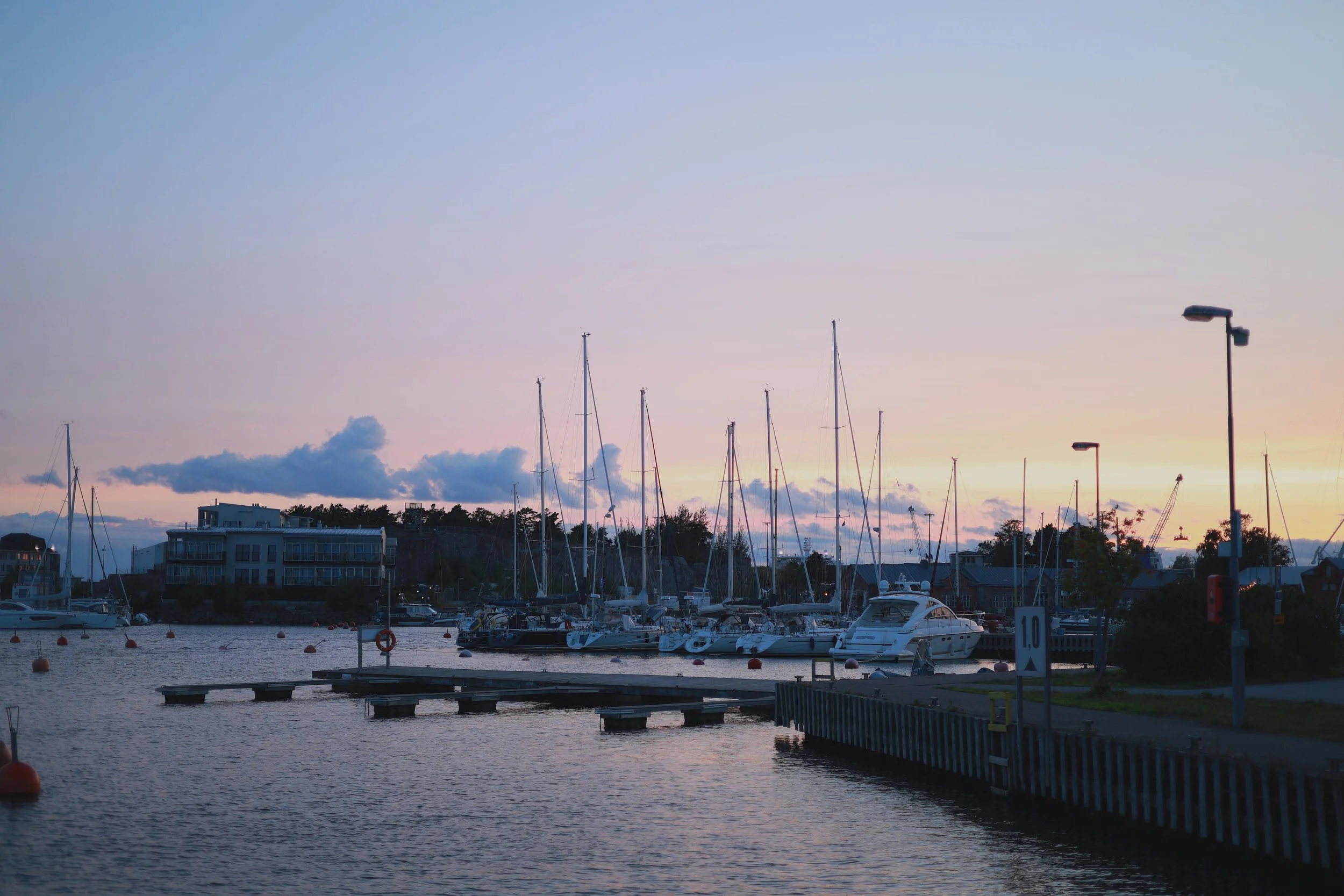 Harbour with boats and sailboat masts beneath a pastel blue and pink evening sky.