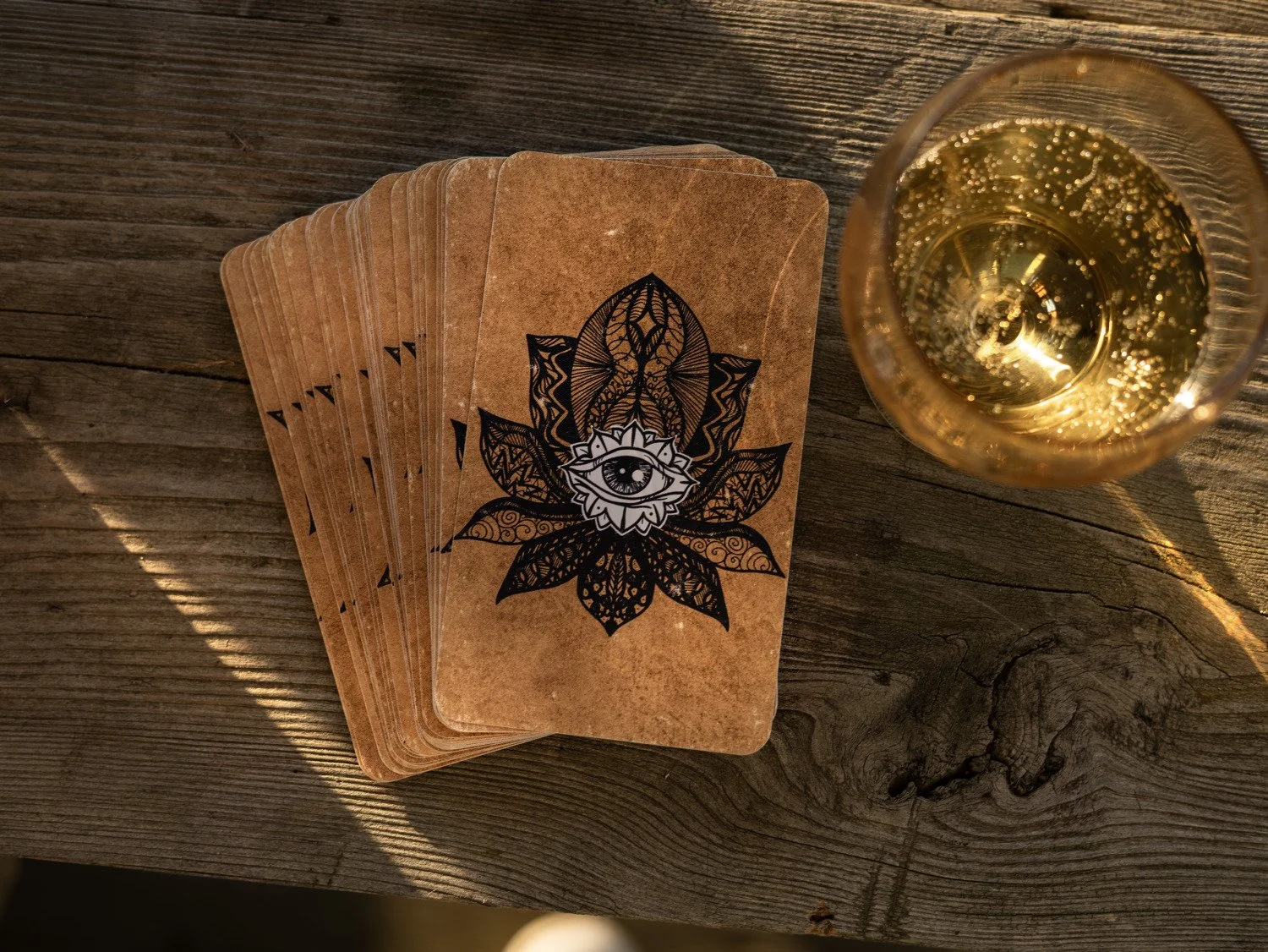 Brown oracle card deck with a black floral eye design beside a glass on a weathered wooden table.