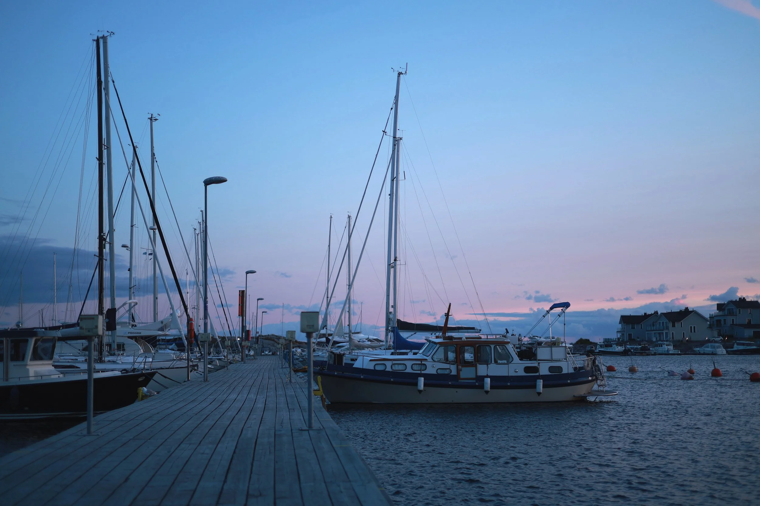 Blue-hour harbour with a wooden pier, boats and tall sailboat masts under a pink horizon.