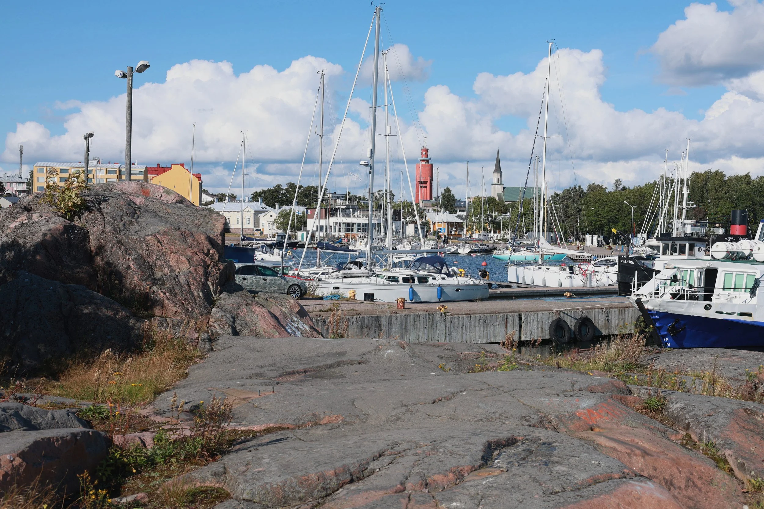 Rocky shoreline leading toward a harbour with sailboats, buildings and a red lighthouse.