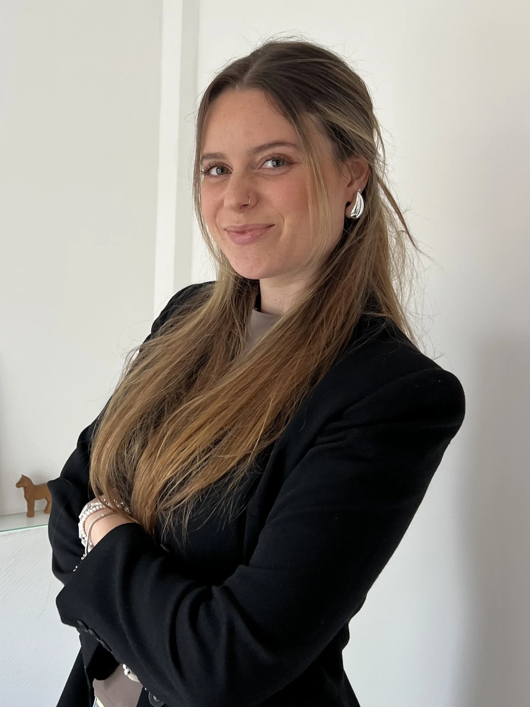 Junge Frau mit langen, braunen Haaren, schwarzer Jacke und silbernen Ohrringen, die vor einer weißen Wand steht und lächelt.