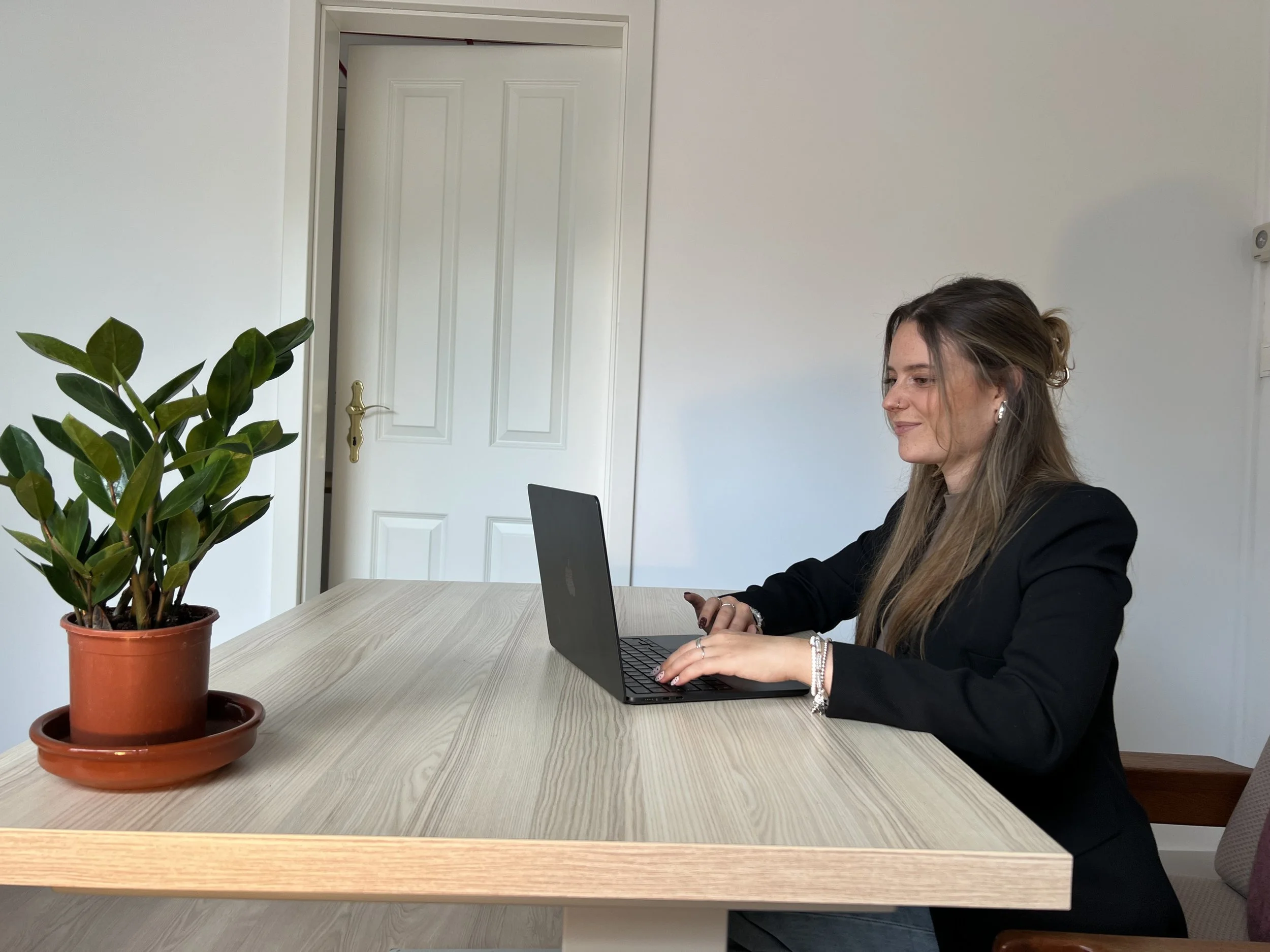 Eine junge Frau sitzt an einem Holztisch, arbeitet an einem Laptop, trägt eine schwarze Jacke und hat braune, wallende Haare. Auf dem Tisch befindet sich eine Topfpflanze in einem braunen Blumentopf.
