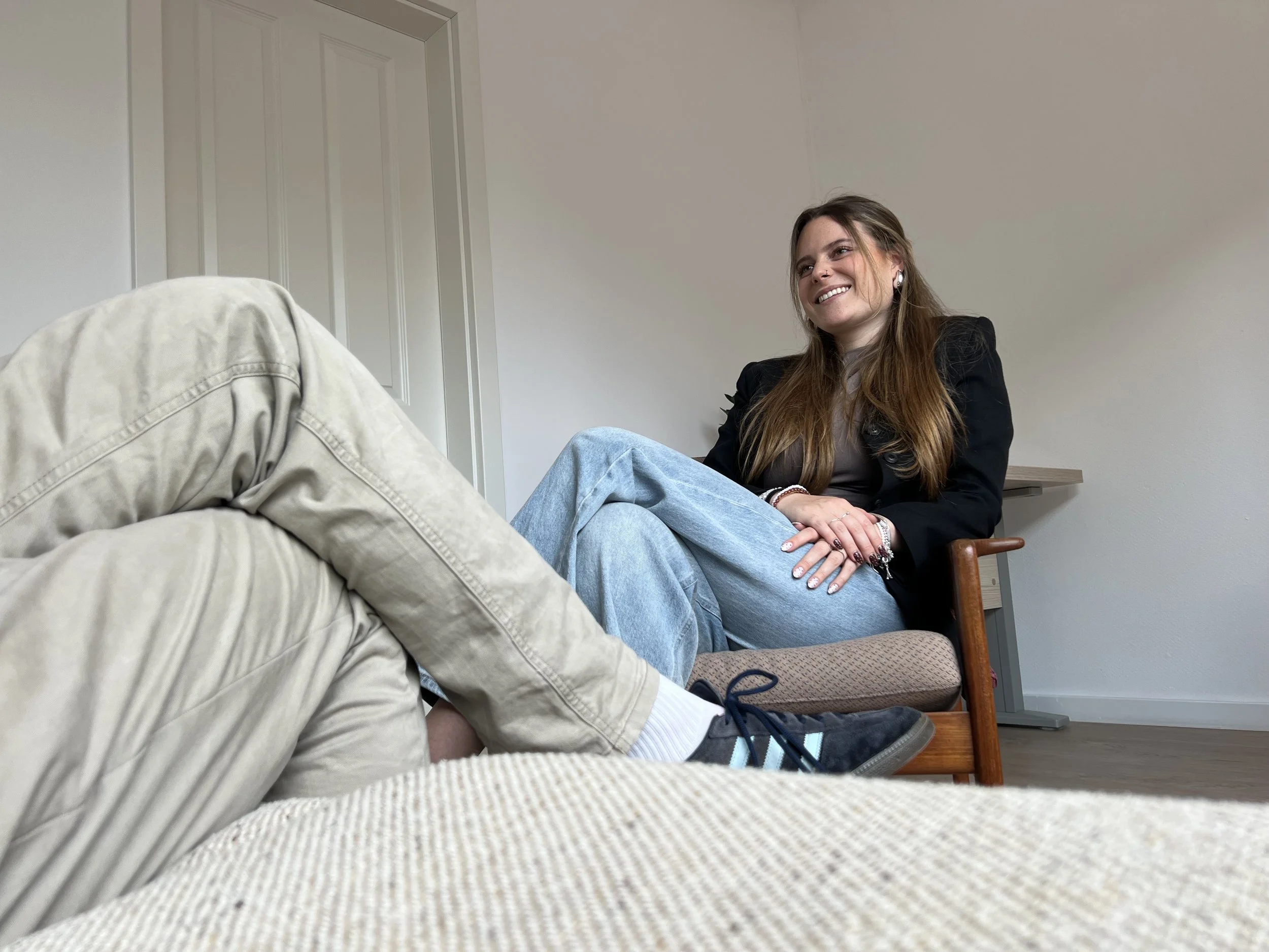 Eine lachende Frau sitzt auf einem Stuhl, während eine Person auf der Couch im Vordergrund liegt. Das Bild zeigt ein Gespräch in einem neutral eingerichteten Raum.