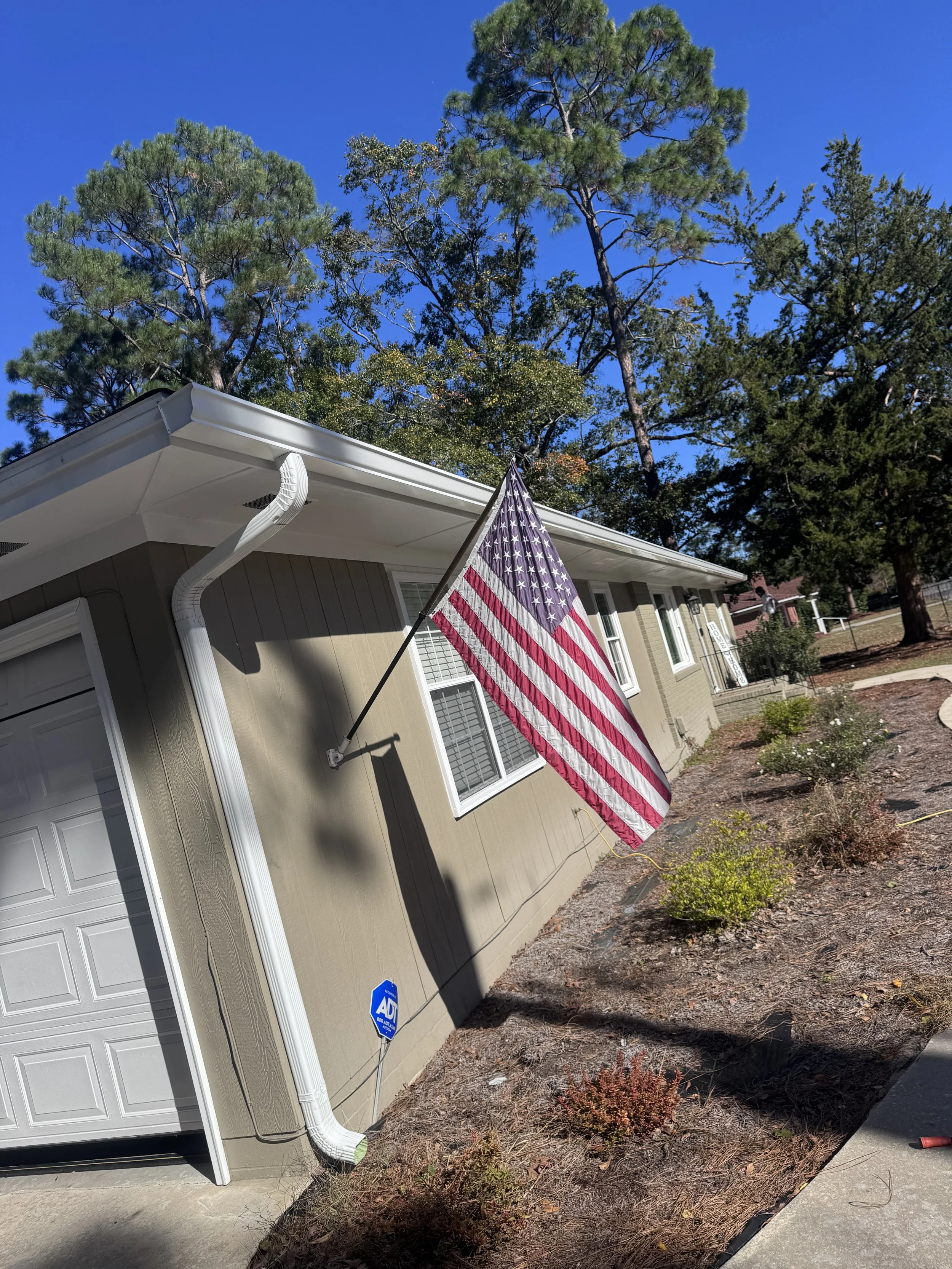 American flag hanging on the side of a house next to a garage, with trees and a blue sky in the background. Gutters Seamless 6" Seamless Gutters
