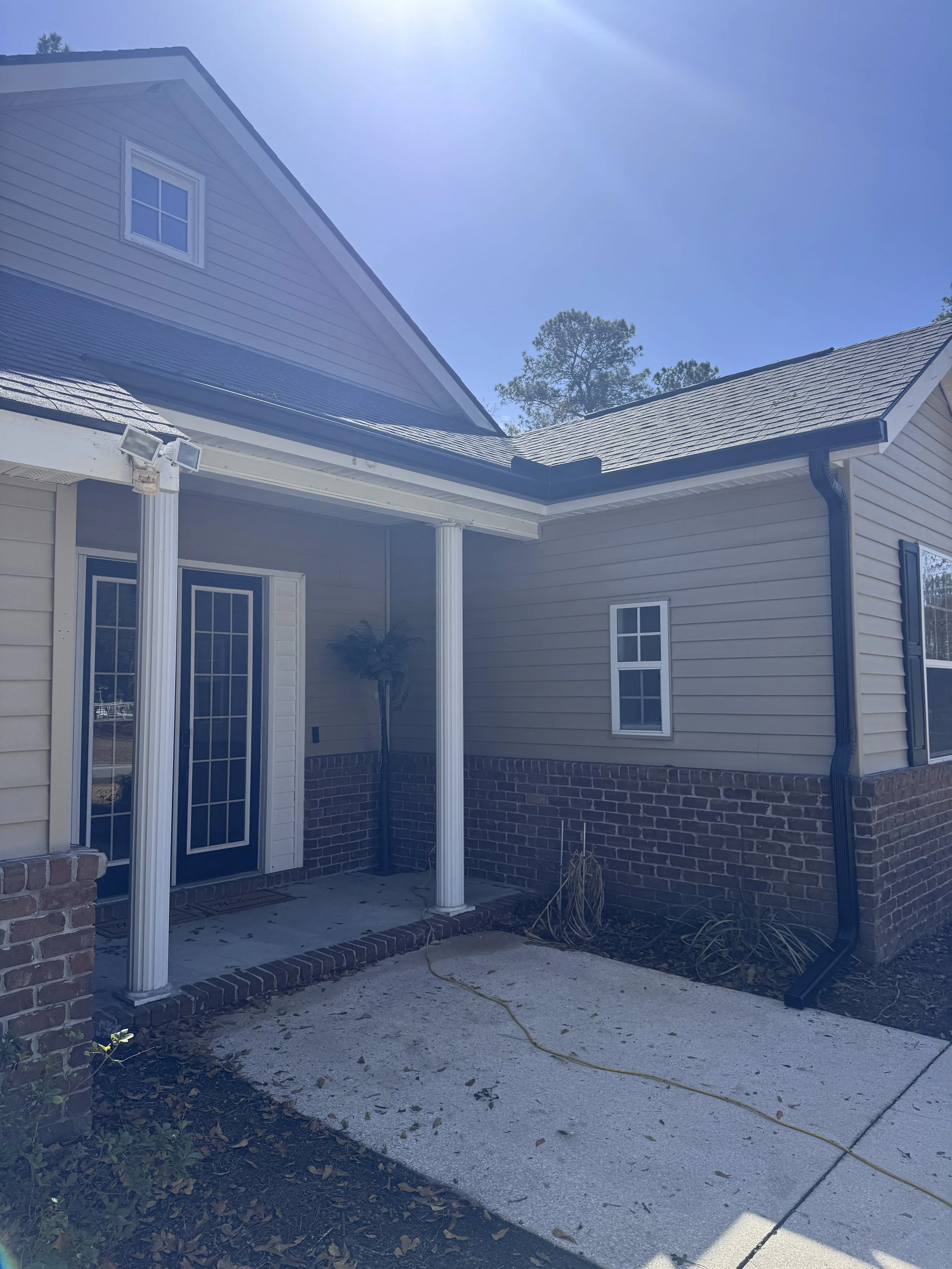 Front porch of a house with two columns, a glass door, a small window, a palm plant, and garden area, under a sunny sky. Gutters Seamless 6" Seamless Gutters