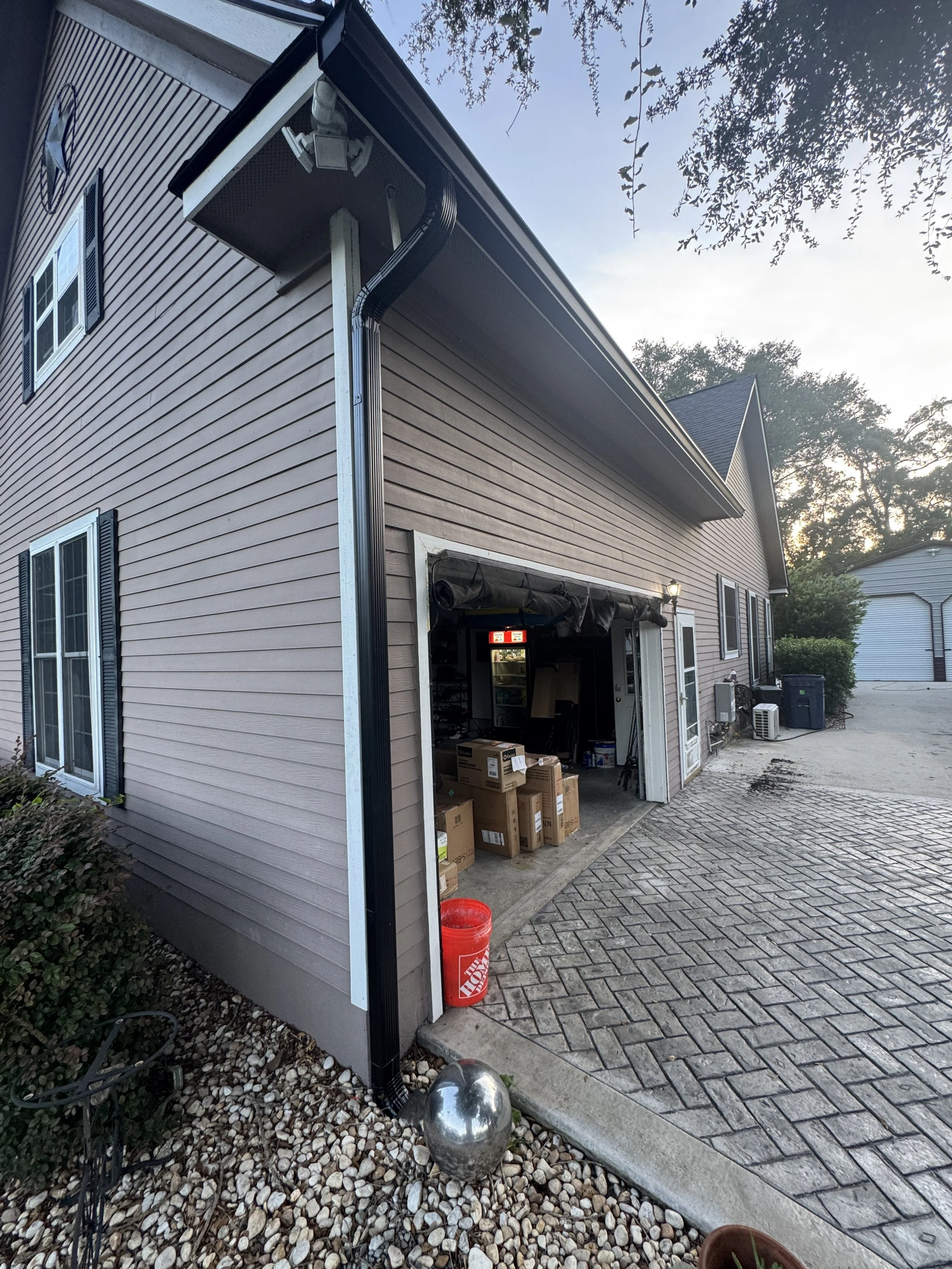 View of a house garage with boxes and supplies inside, outdoor paved driveway, and decorative metal ball on rocks in front. Gutters Seamless 6" Seamless Gutters