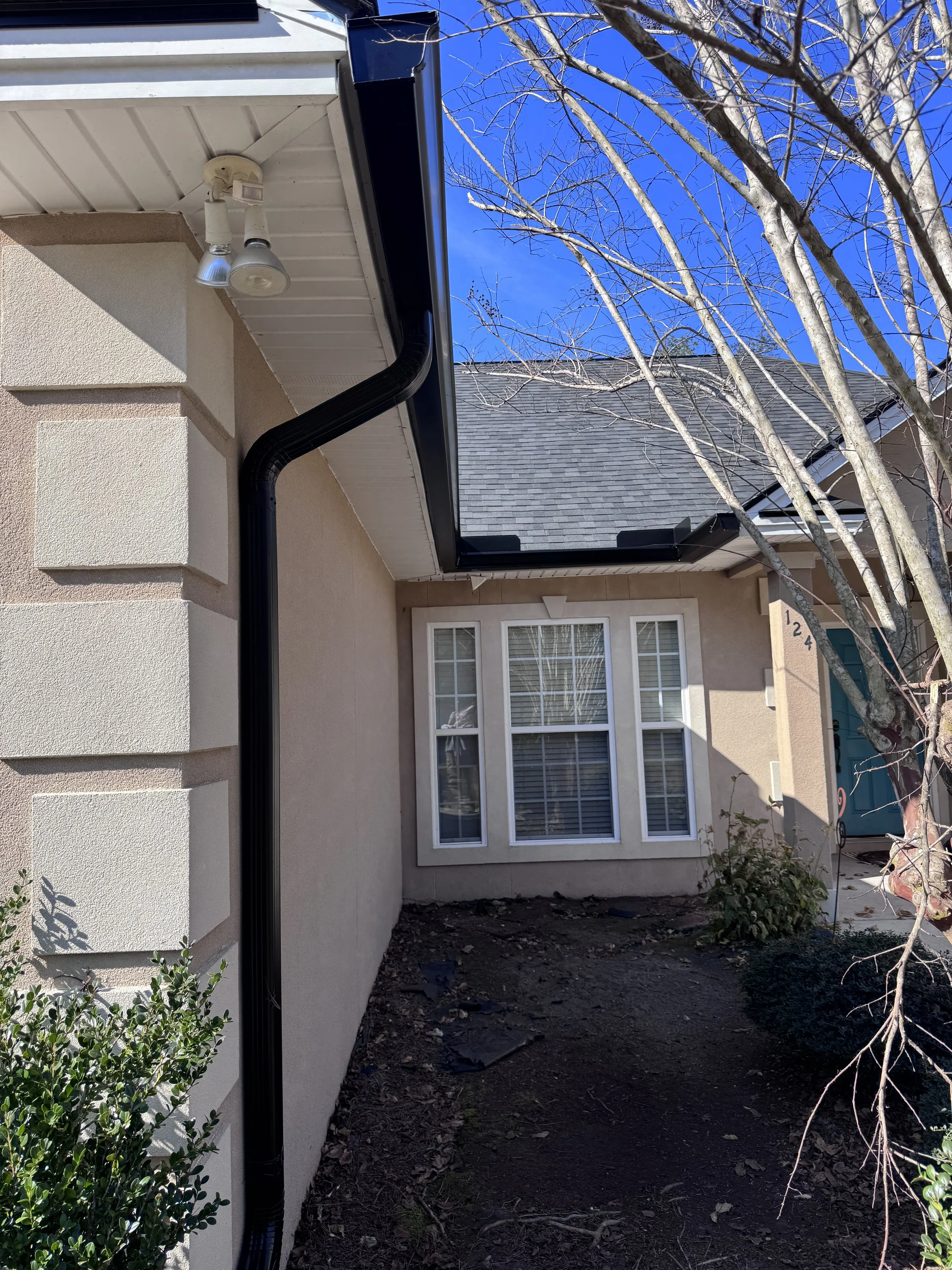 Front view of a house with a black gutter and downspout, beige stucco exterior, a large window with white trim, a tree with no leaves, and a bright blue sky. Gutters Seamless 6" Seamless Gutters
