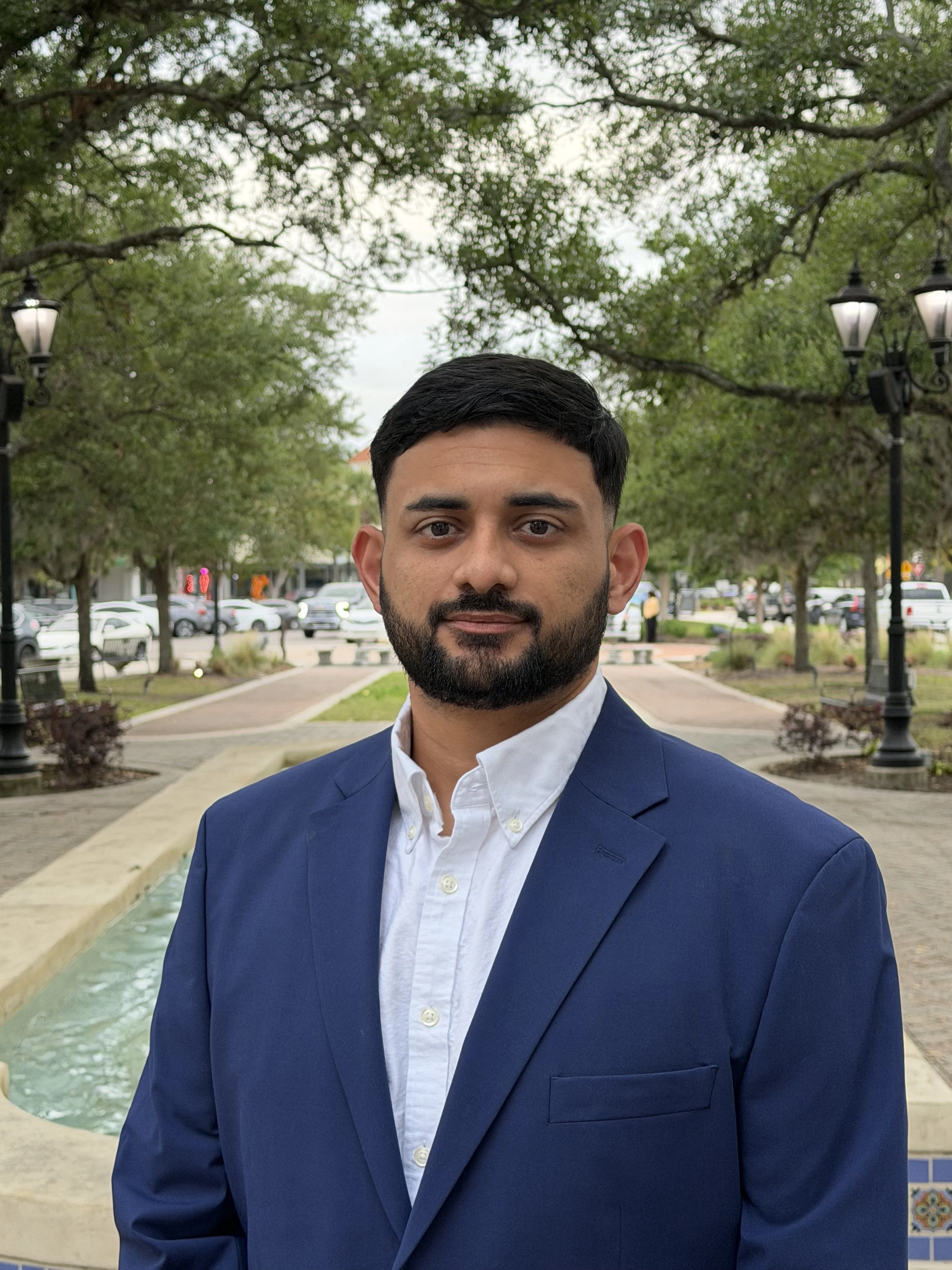 A man in a blue suit and white shirt standing outdoors in a park with trees, street lamps, and parked cars in the background. Florida Insurance Erik Sifuentes Insurance Auto Insurance Home Insurance Business Insurance Erik Sifuentes Licensed Agent