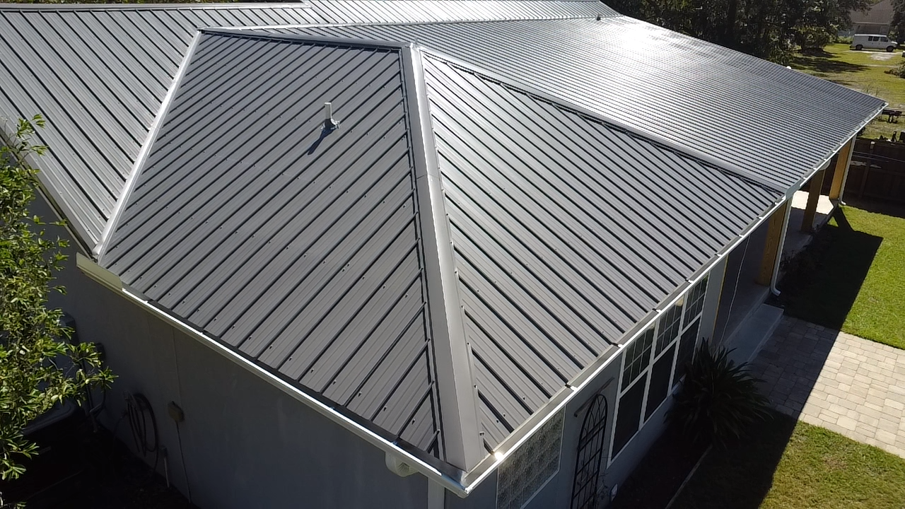 A house with a gray metal roof, showing both the larger sloped section and a smaller flat section with a vent pipe. Gutters Seamless 6" Seamless Gutters