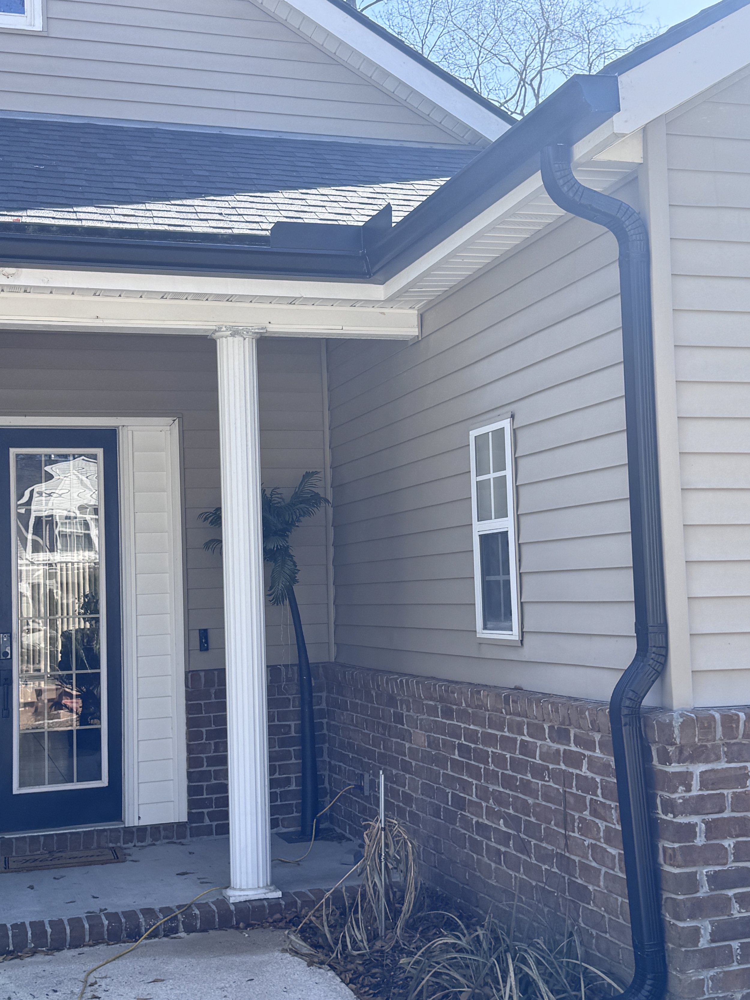 Exterior view of a house showing a corner with brick and beige siding, a white column, a window, a door, and a black downspout. Gutters Seamless 6" Seamless Gutters