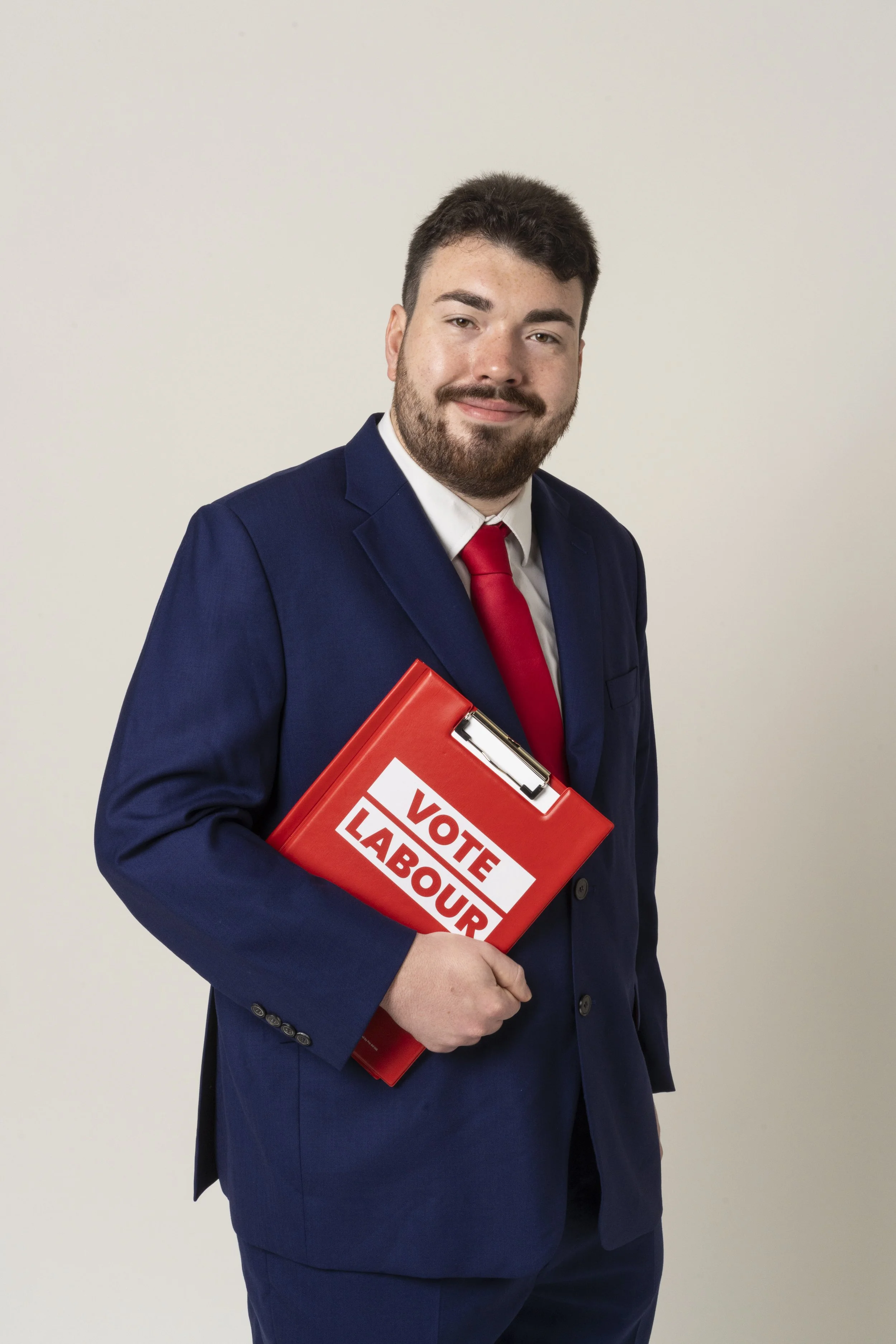 A young man in a navy blue suit, white shirt, and red tie, holding a red folder, standing against a gray background.