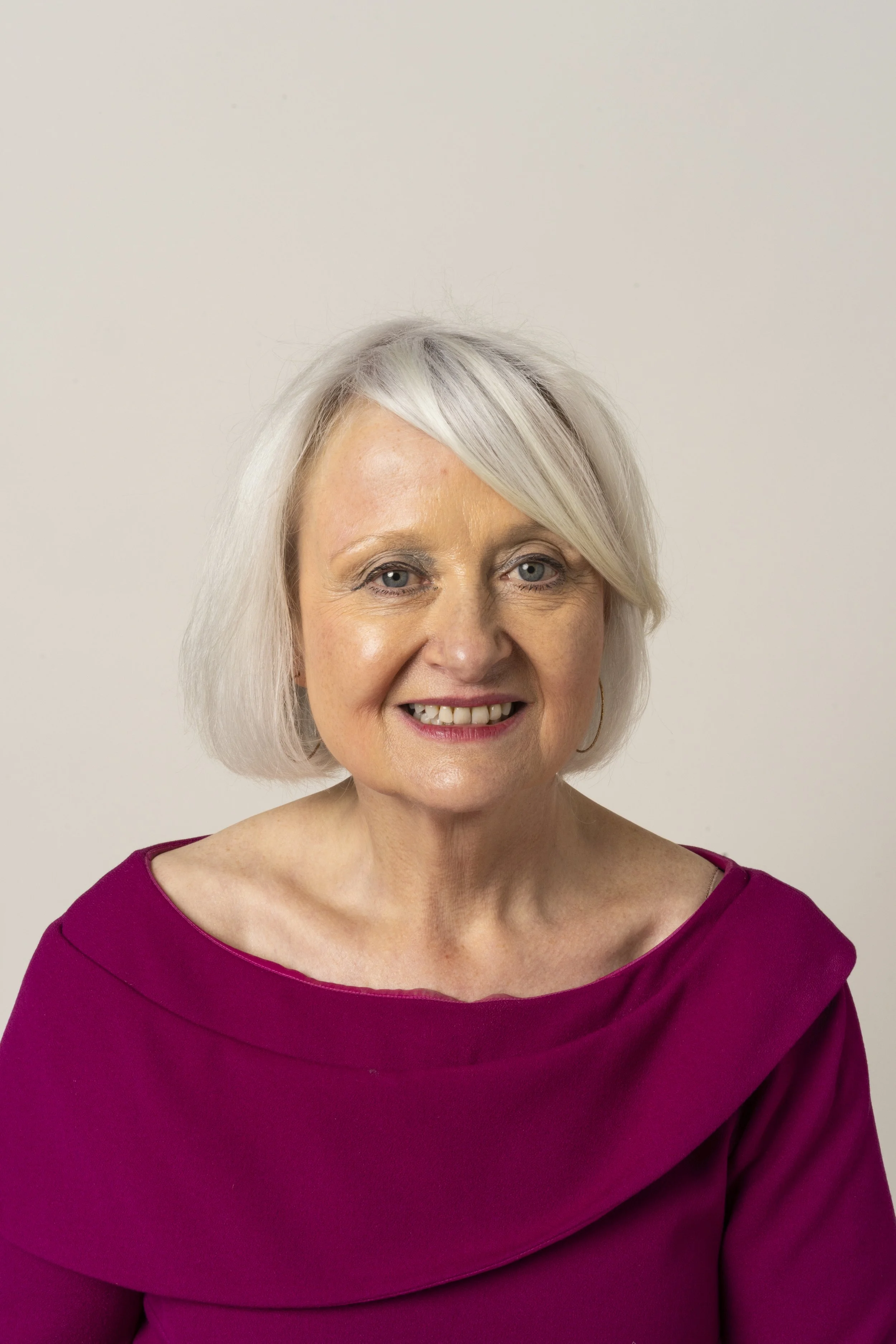 A woman with short, grey hair, wearing a black top, pearl earrings, and a layered pearl necklace, against a gray background.