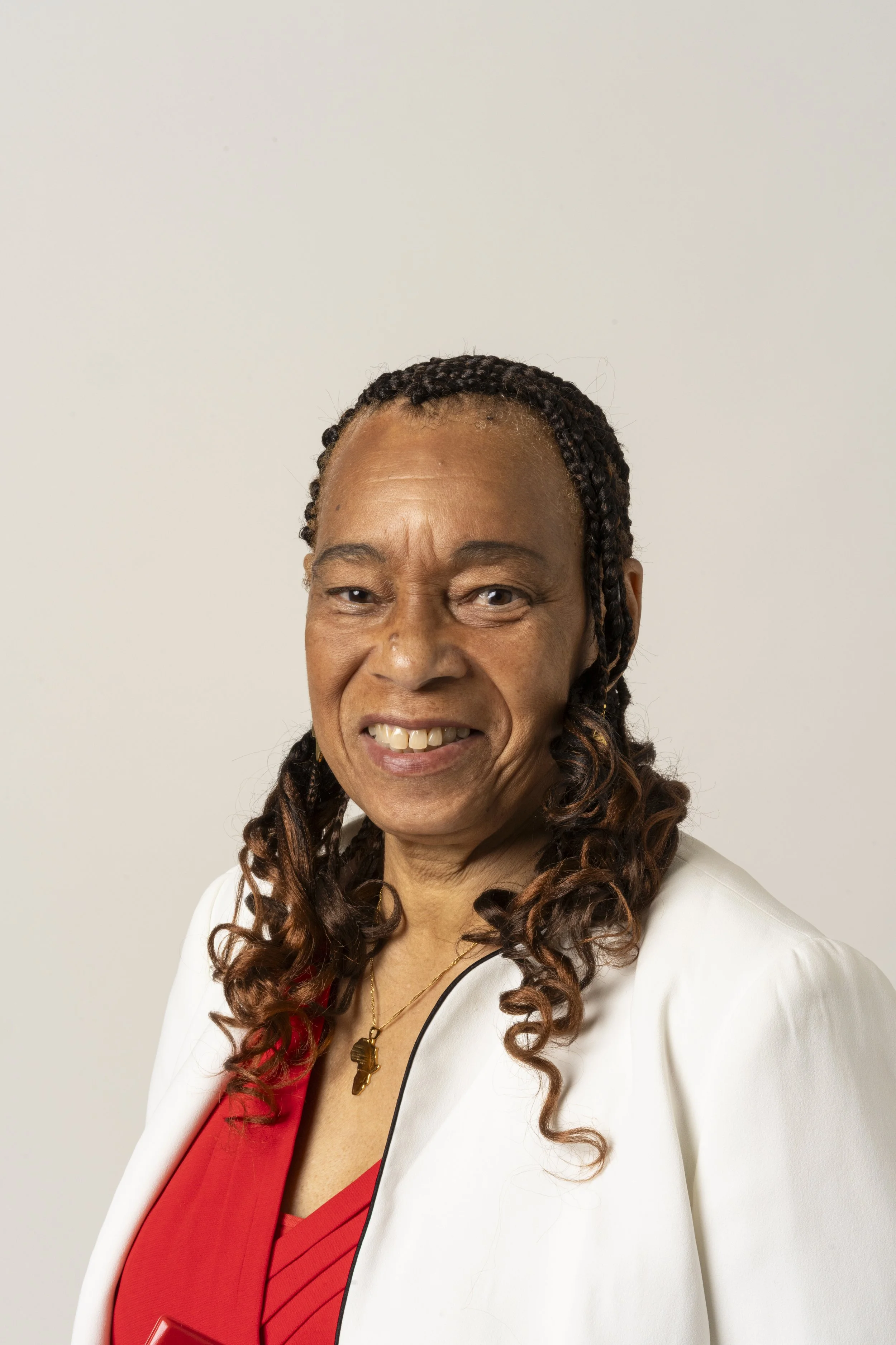 Portrait of a smiling woman with braids, wearing a white blazer and a red lace top, against a plain, light background.
