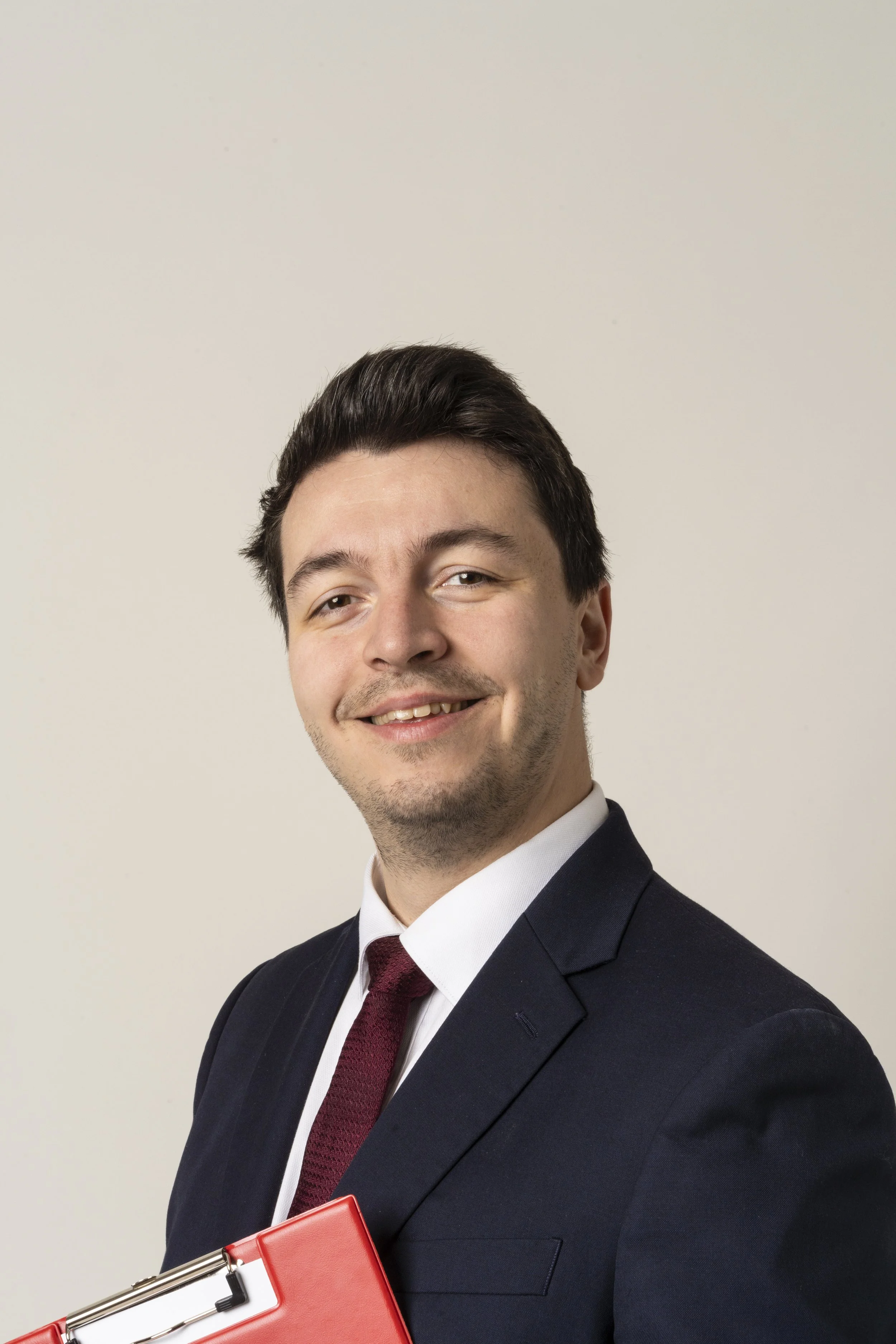 A young man in a suit and tie smiling at the camera, holding a red folder.